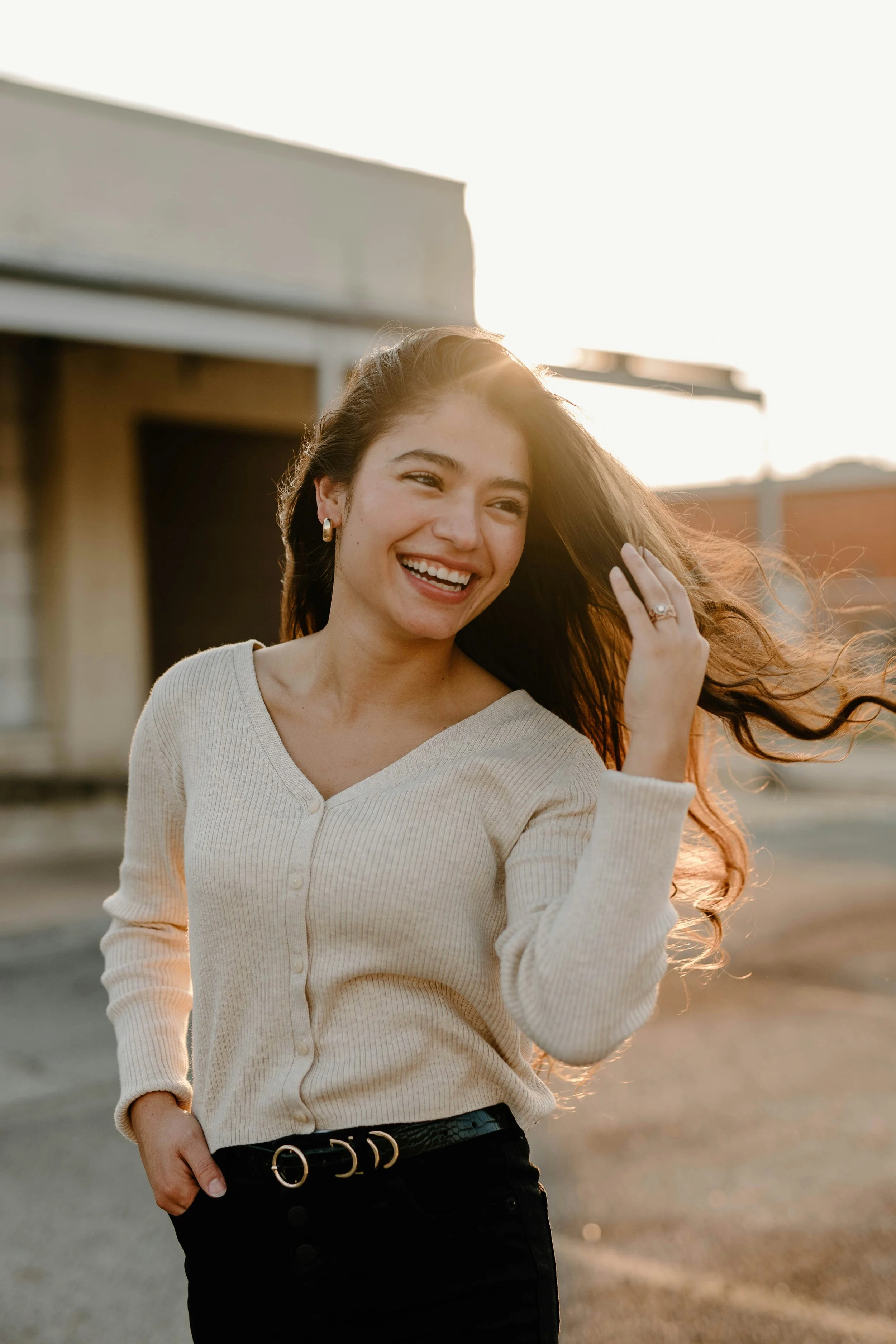 A young woman with long hair, smiling and touching her hair, standing outdoors near a building during sunset. She is the perfect representation of the kind of teen patients seen at the Bend Oregon Direct Primary Care office of Alpine Wellness
