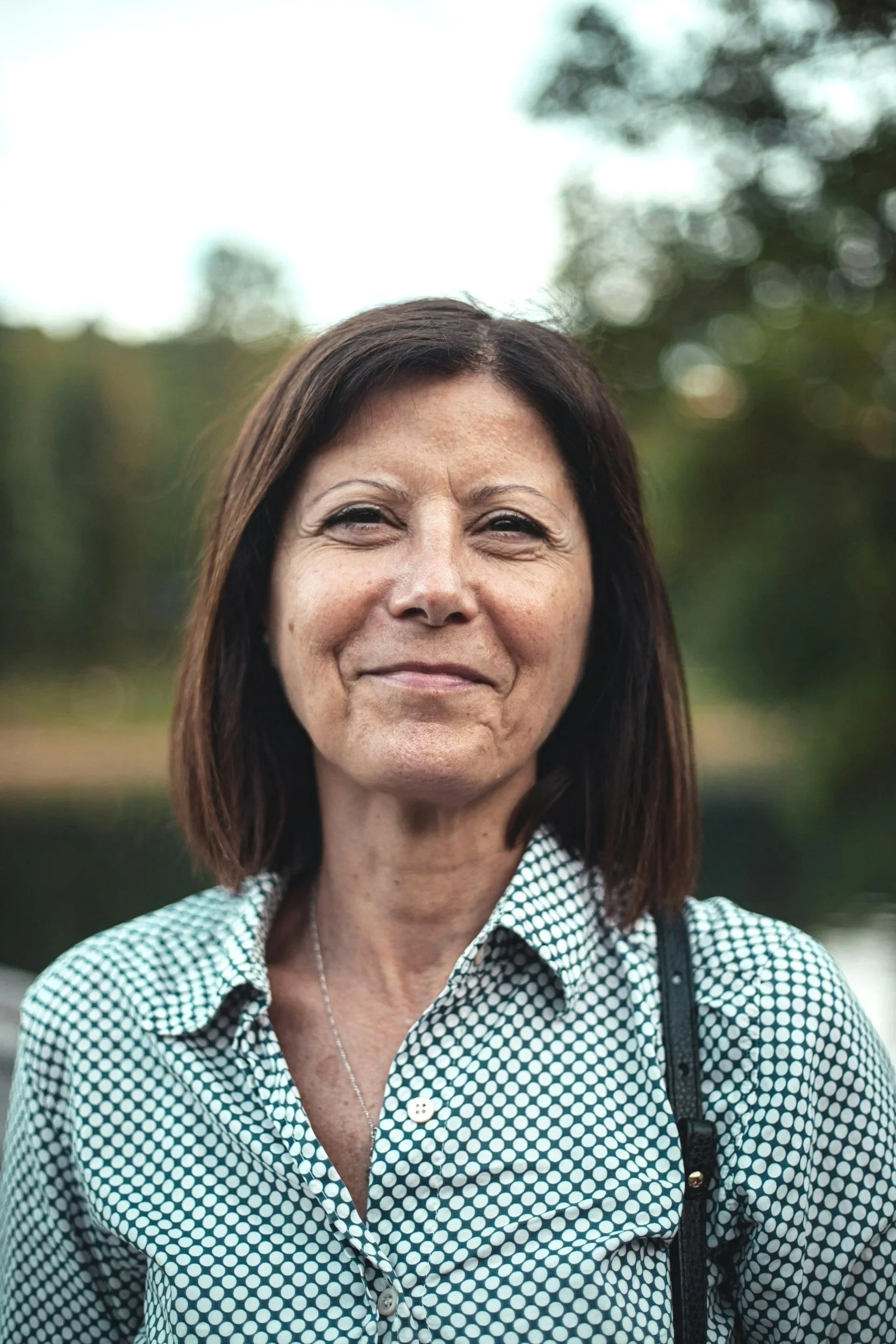 Close-up of a woman with shoulder-length brown hair, wearing a black and white checkered shirt, outdoors with blurred trees in the background, she is looking forward to meeting with her hormone doctor in central Oregon