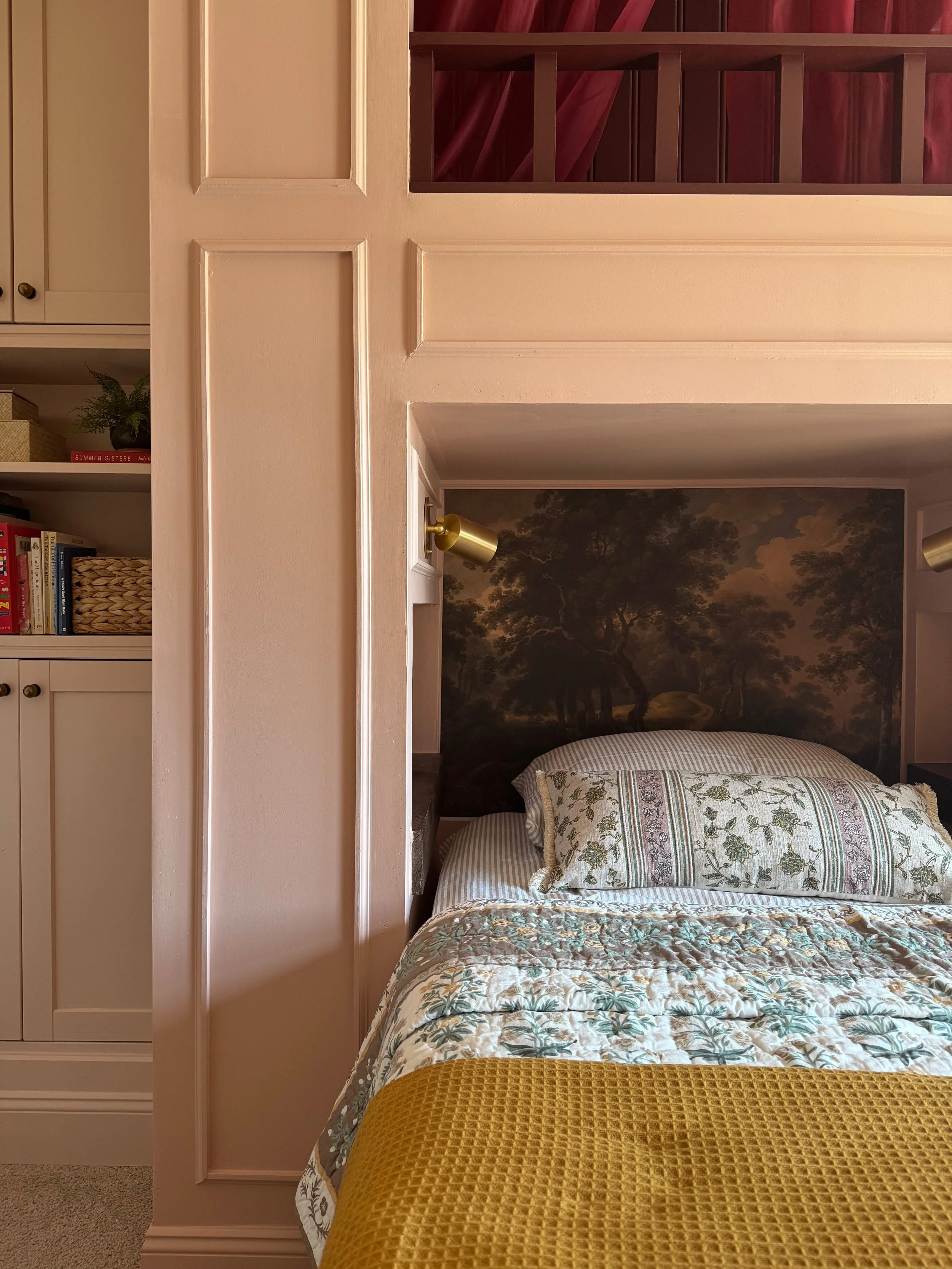 A bedroom with a reversed view of the bed, featuring a patterned pillow, a bedspread with floral designs, and a yellow blanket. The headboard wall has a tree mural, and the room has two gold wall-mounted reading lights. There are built-in white shelves and cabinets to the left, with books and decorative items.