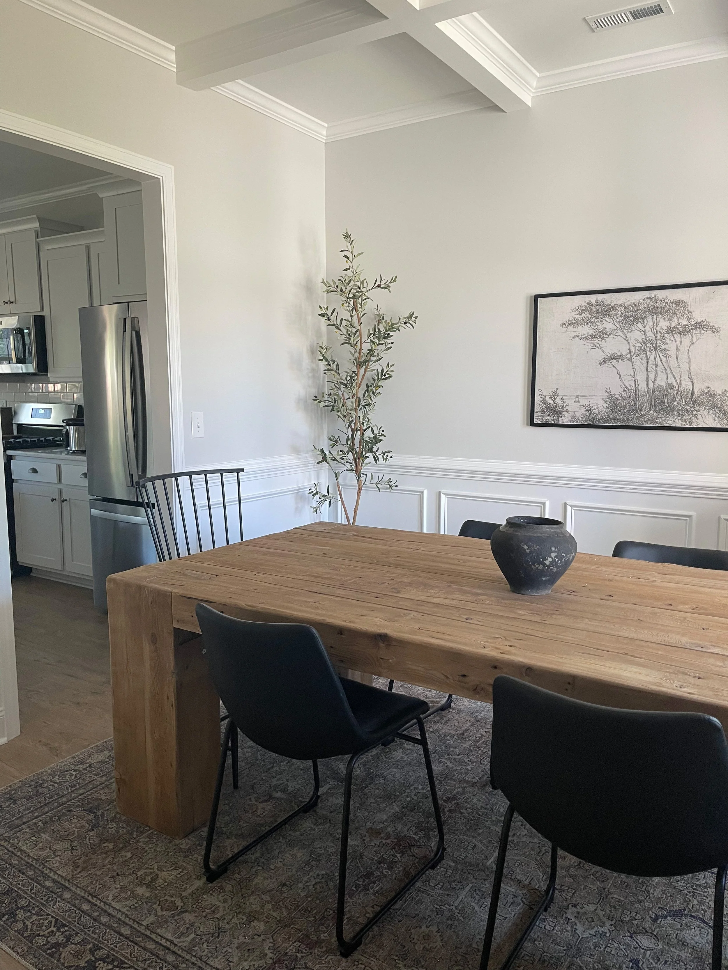 A dining room with a wooden table, black chairs, a black vase on the table, a potted plant in the corner, and a framed sketch of trees on the wall.