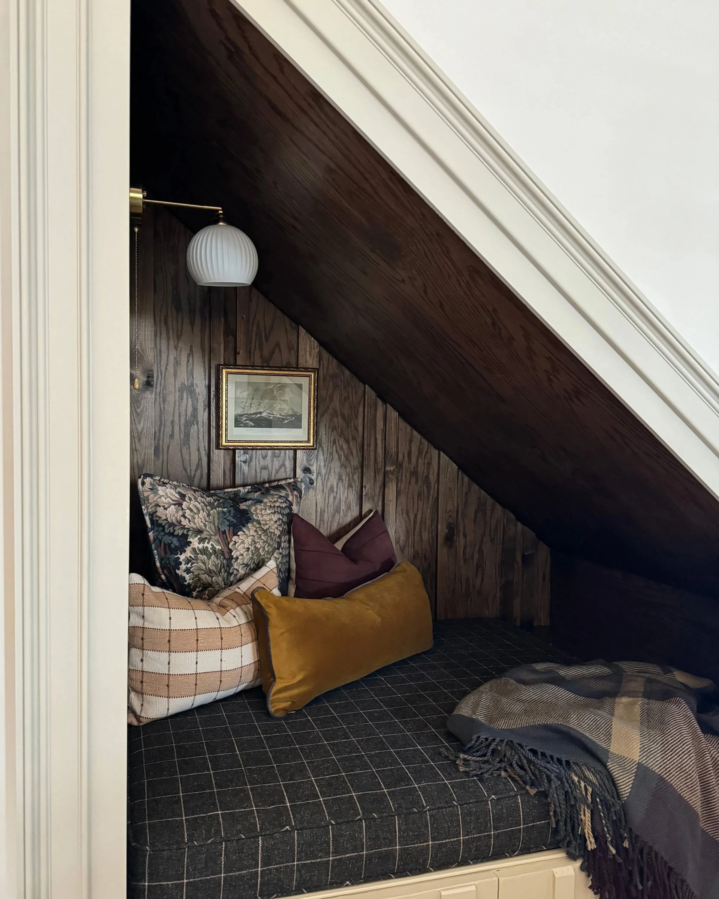 Cozy nook under angled dark wood ceiling with pillows, a framed picture, a wall-mounted lamp, and a plaid blanket.