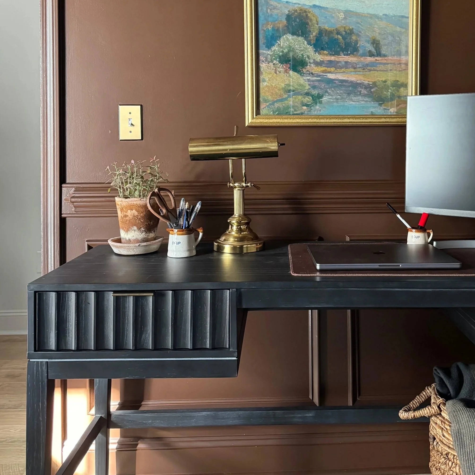 A black desk with a potted plant, a brass desk lamp, a small cup holding pens and scissors, and a computer monitor in front of a brown wall with a landscape painting.