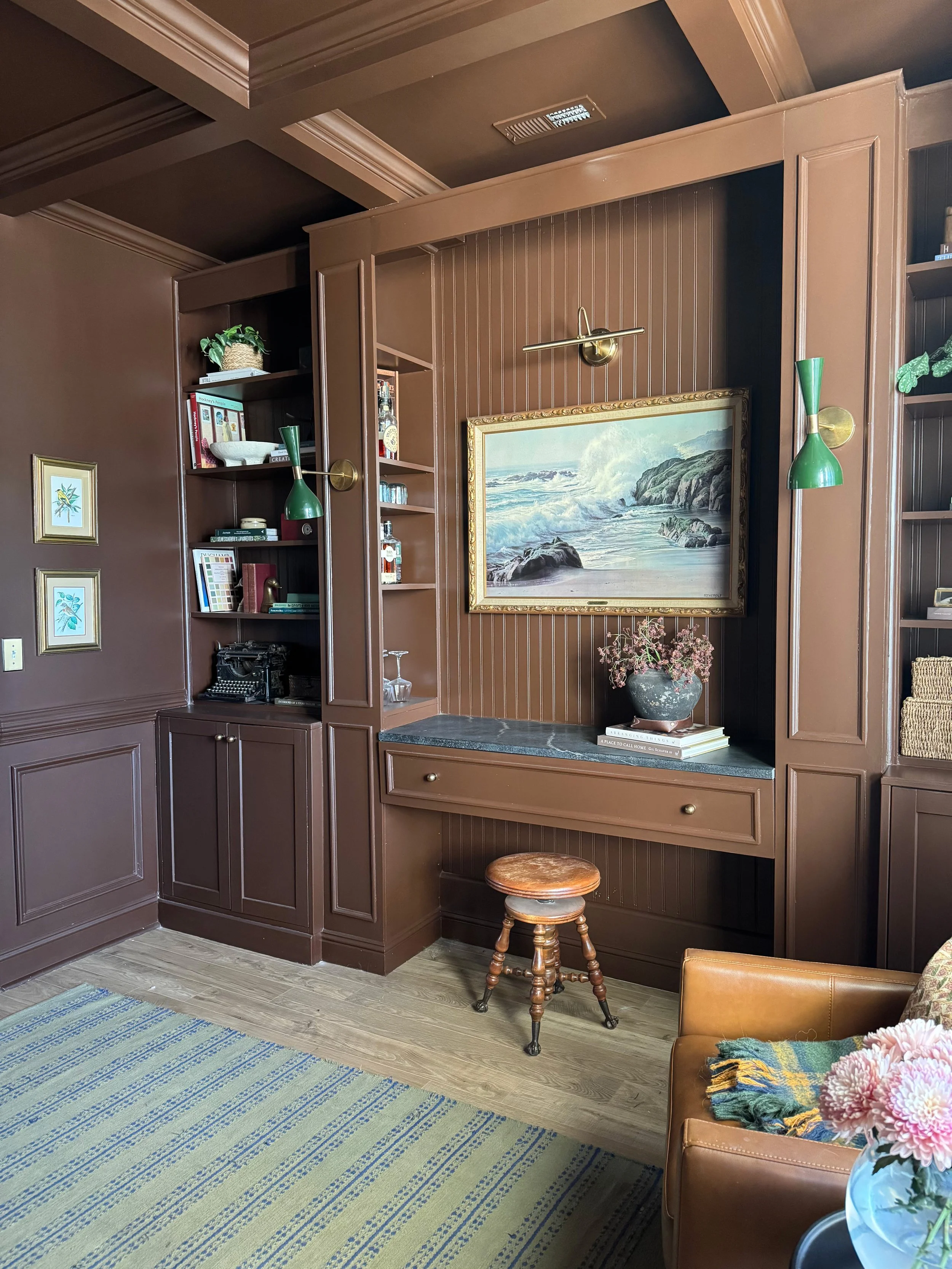 A built-in wall unit with shelves and drawers, decorated with a seaside painting, a potted plant, and vintage glassware, in a brown-room interior with a patterned Rug and a leather armchair with a blanket.