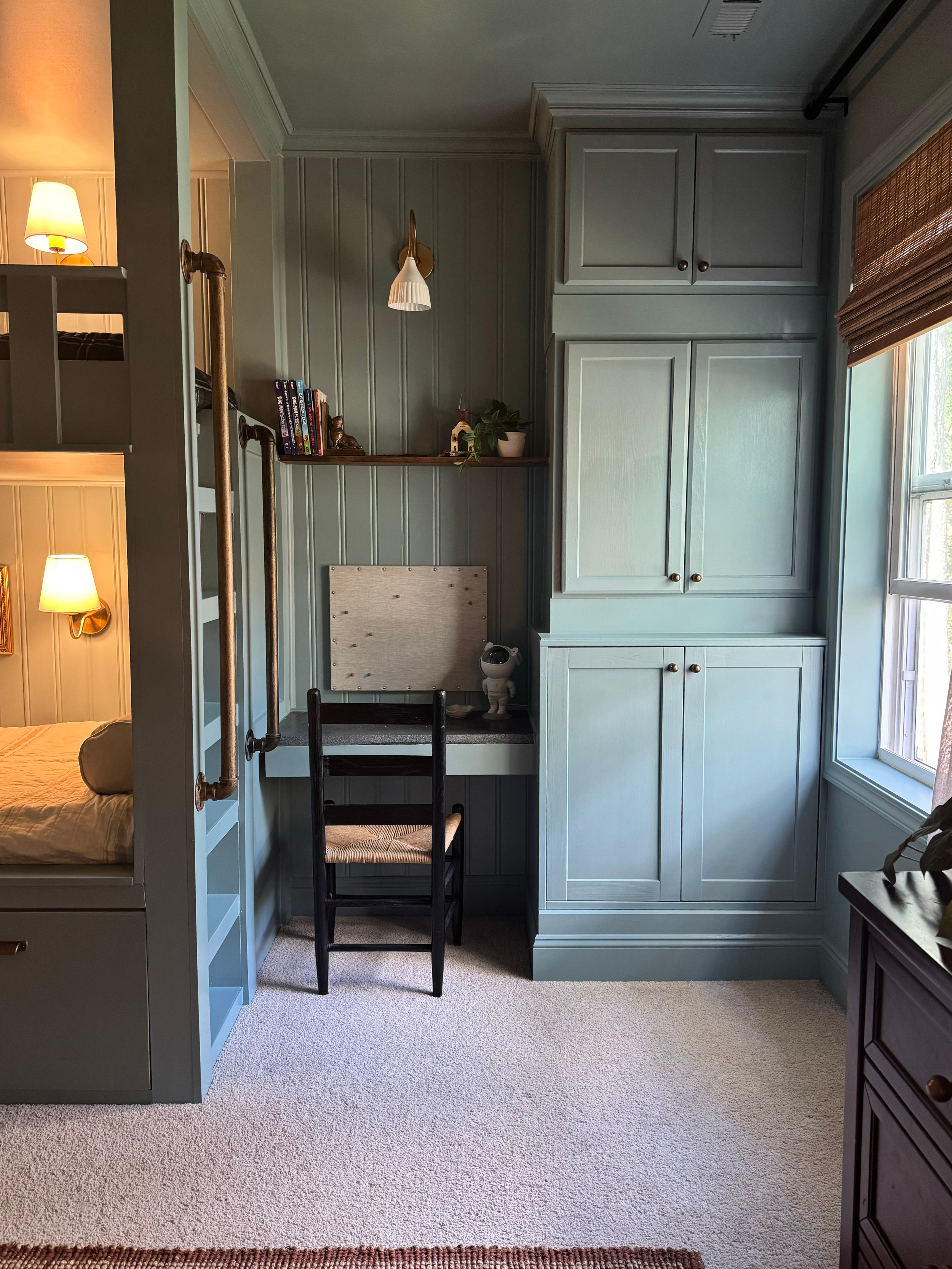 A cozy corner in a bedroom with a built-in light blue desk and cabinetry, a small chair, and decorative items on the shelves. The room has a large window with wooden blinds, and part of a bed with a pillow is visible on the left.