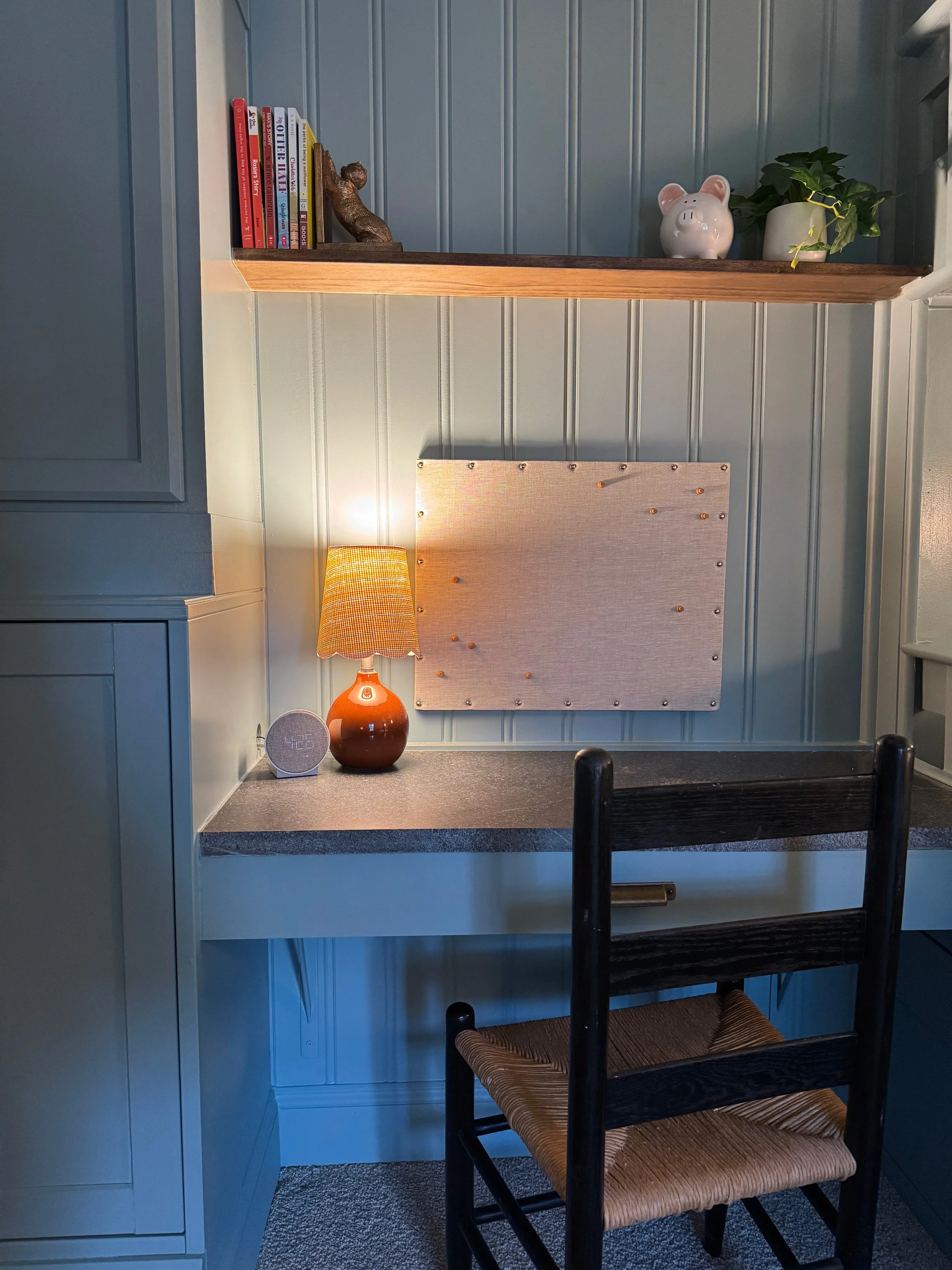 A small workspace with a black desk, a wooden chair with a woven seat, a table lamp, a smart speaker, a corkboard, and decorative items on a shelf above.