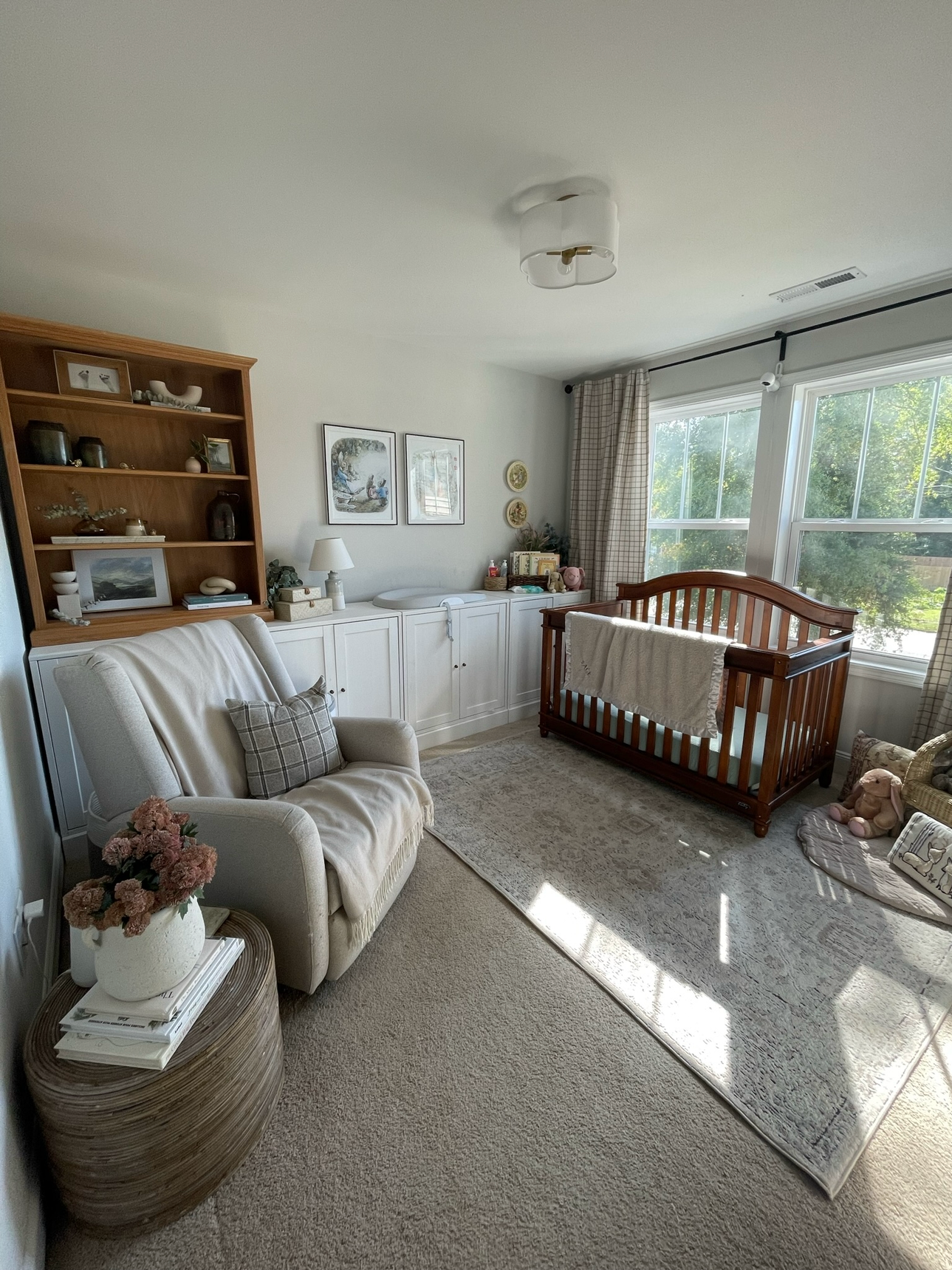 Living room with beige carpet, white armchair with plaid pillow, wooden crib, bookshelf with decorative items, white cabinet, framed artwork on wall, table with vase of pink flowers, window with curtains, sunlight streaming in.