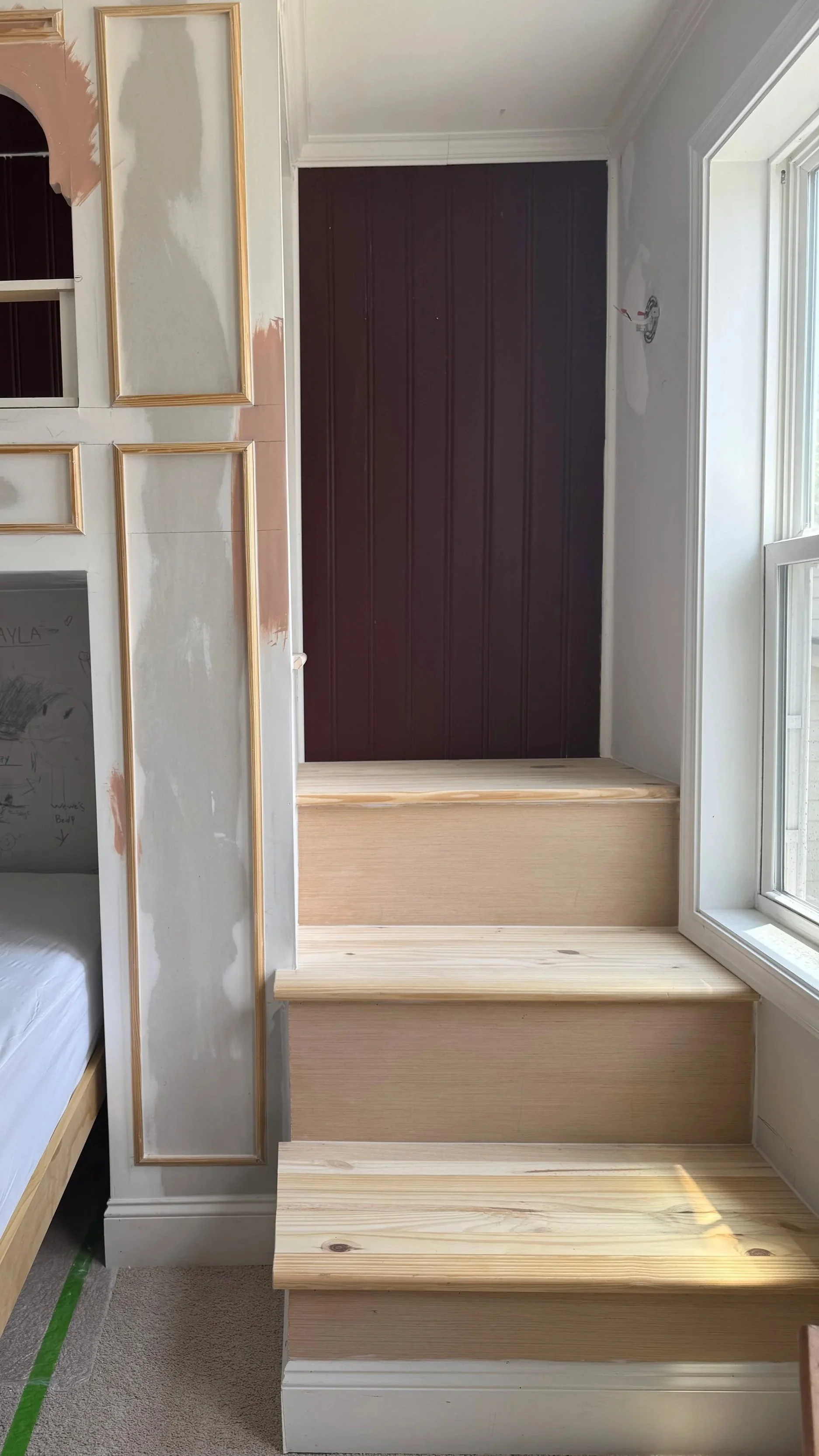 Unfinished staircase with three wooden steps leading up to a dark purple wall, adjacent to a window on the right, and partially painted wall on the left.