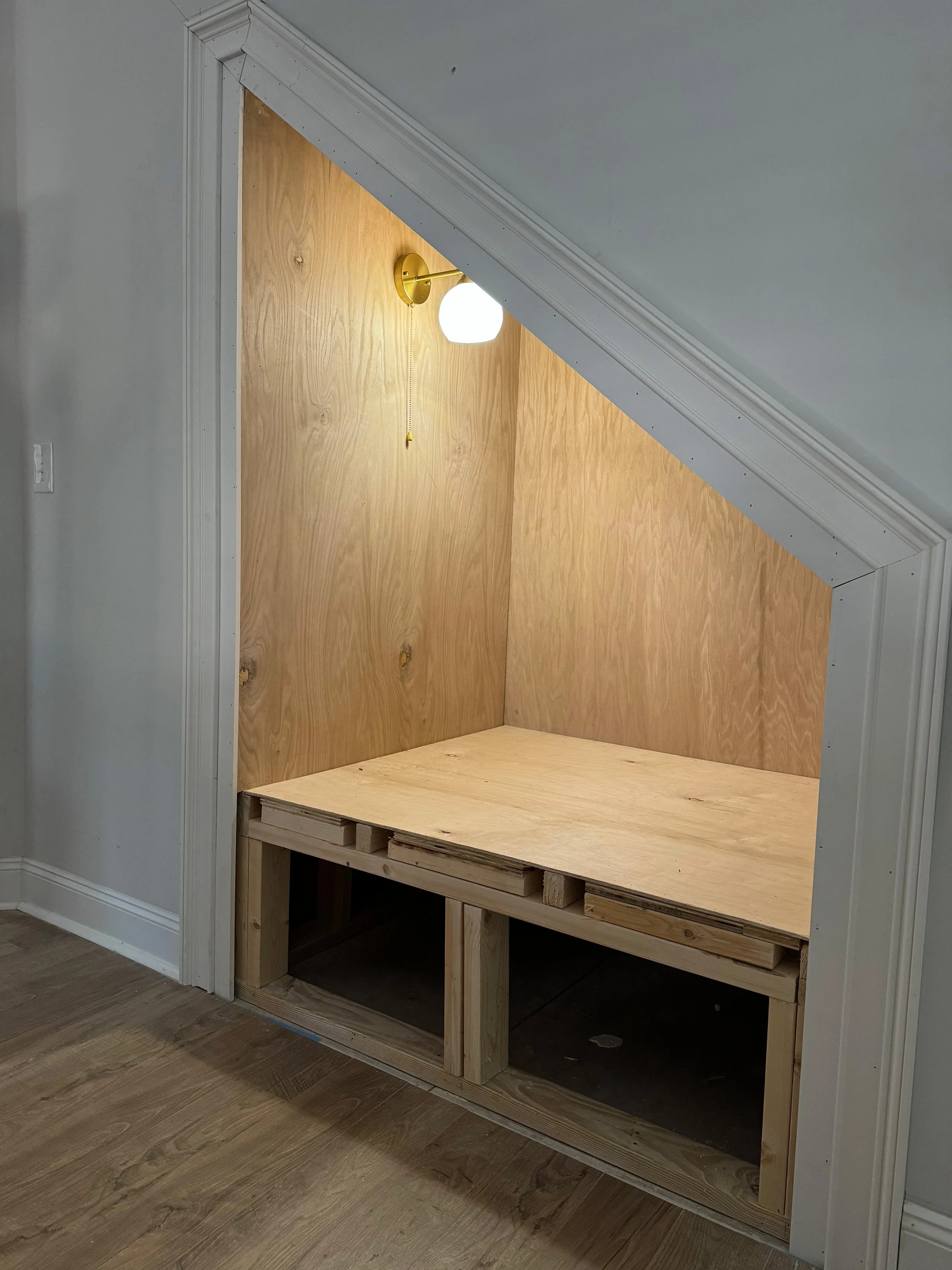 Under-stair built-in nook with plywood backing, a mounted white globe light, and an unfinished wooden platform at the base.