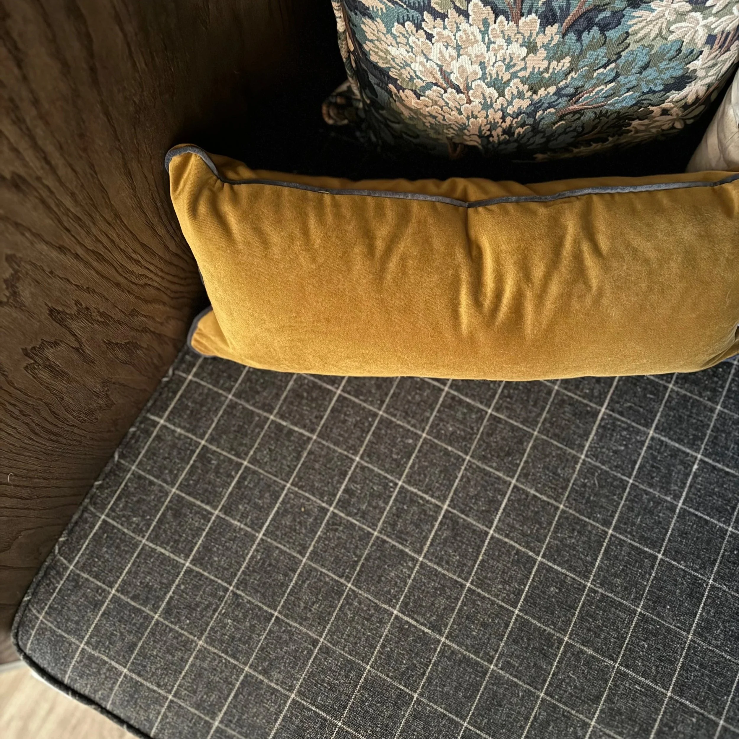 Close-up of a mustard yellow cushion on a gray checkered upholstered bench, with a wooden wall on the left and patterned pillow in the background.