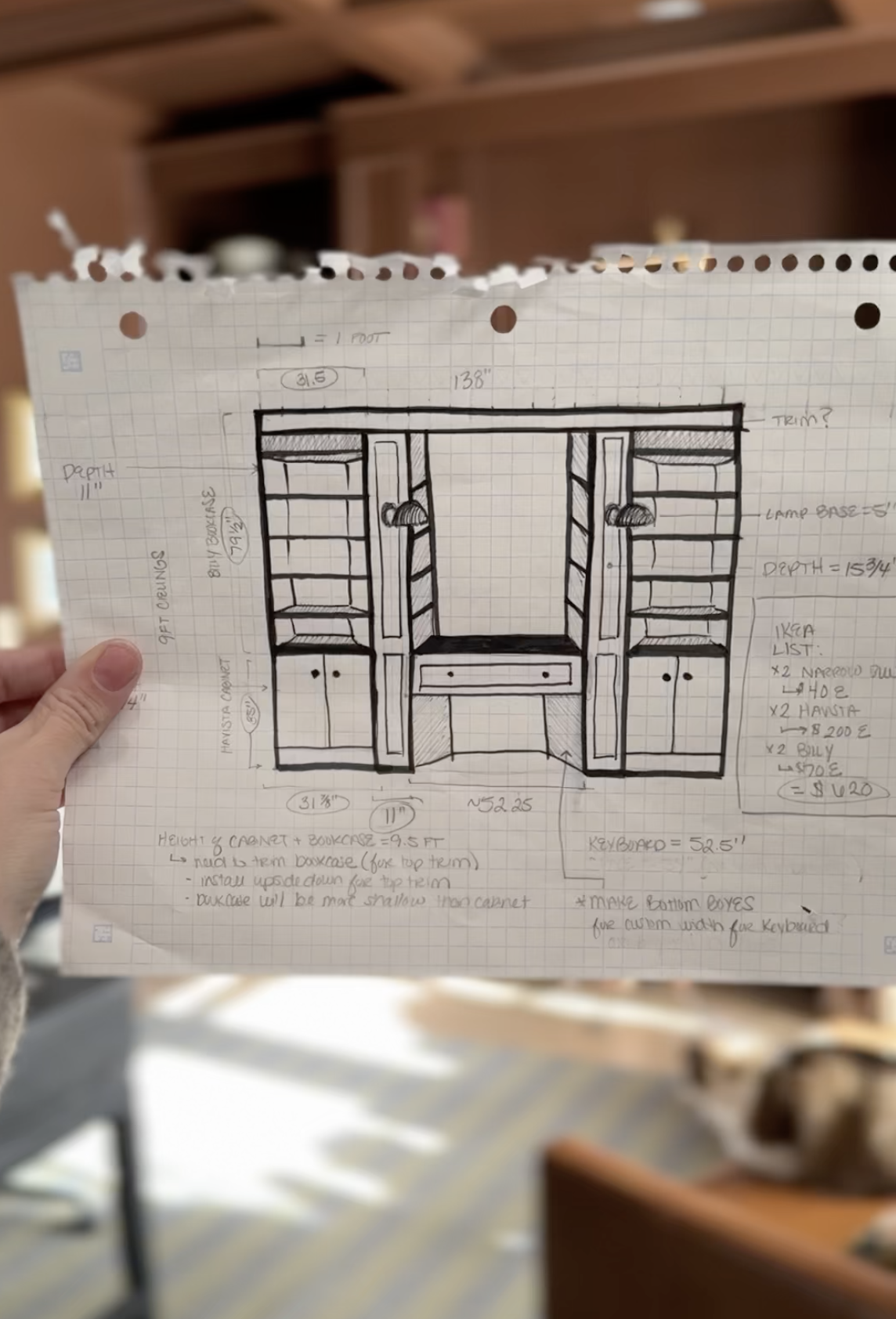 Hand holding a sketch of a built-in shelving unit with dimensions and notes, including shelves, drawers, and a central space.