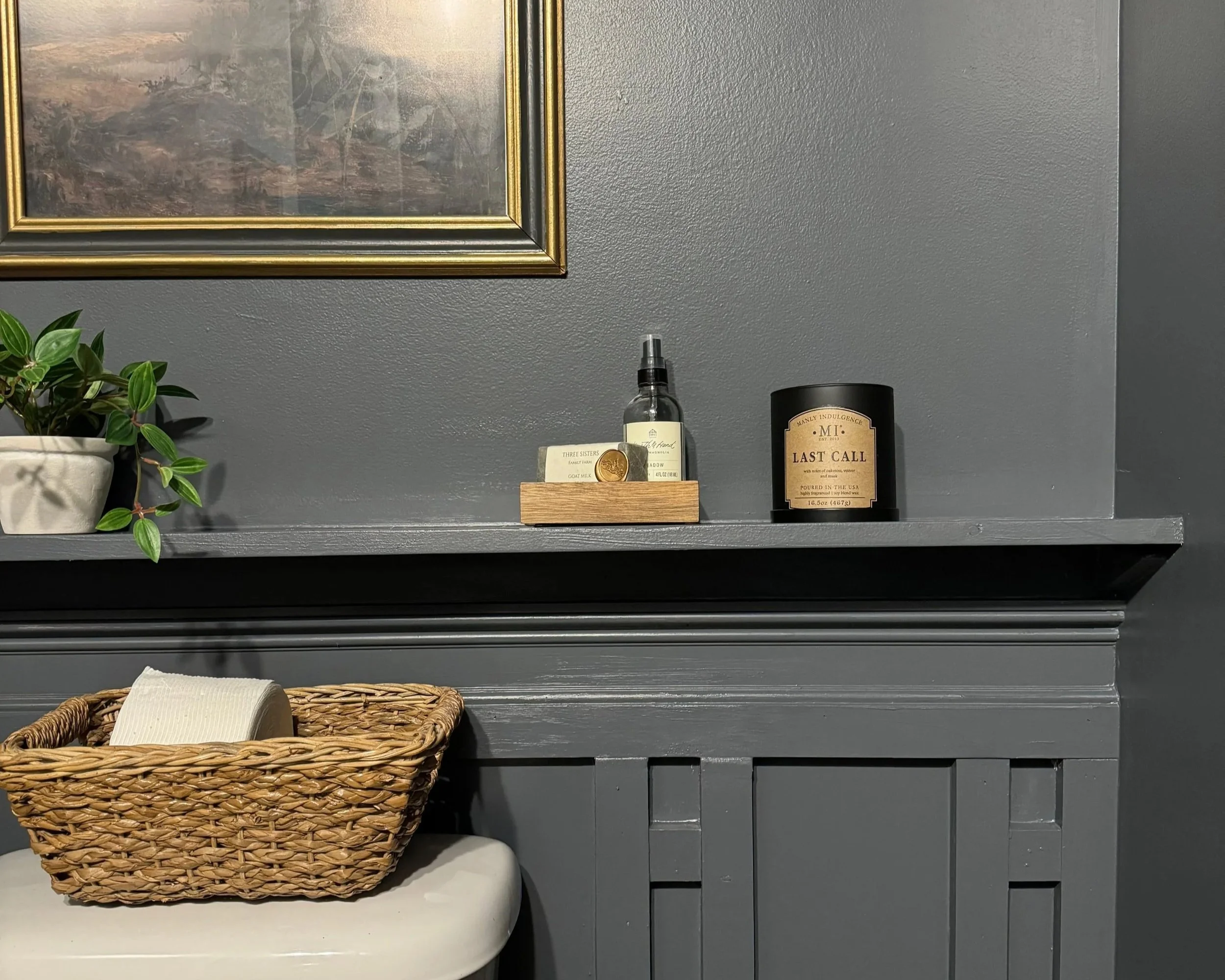 A gray countertop with a small potted green plant, a wooden tray holding a spray bottle, business cards, and a coin, and a black candle labeled 'Last Call' on a dark gray wall.