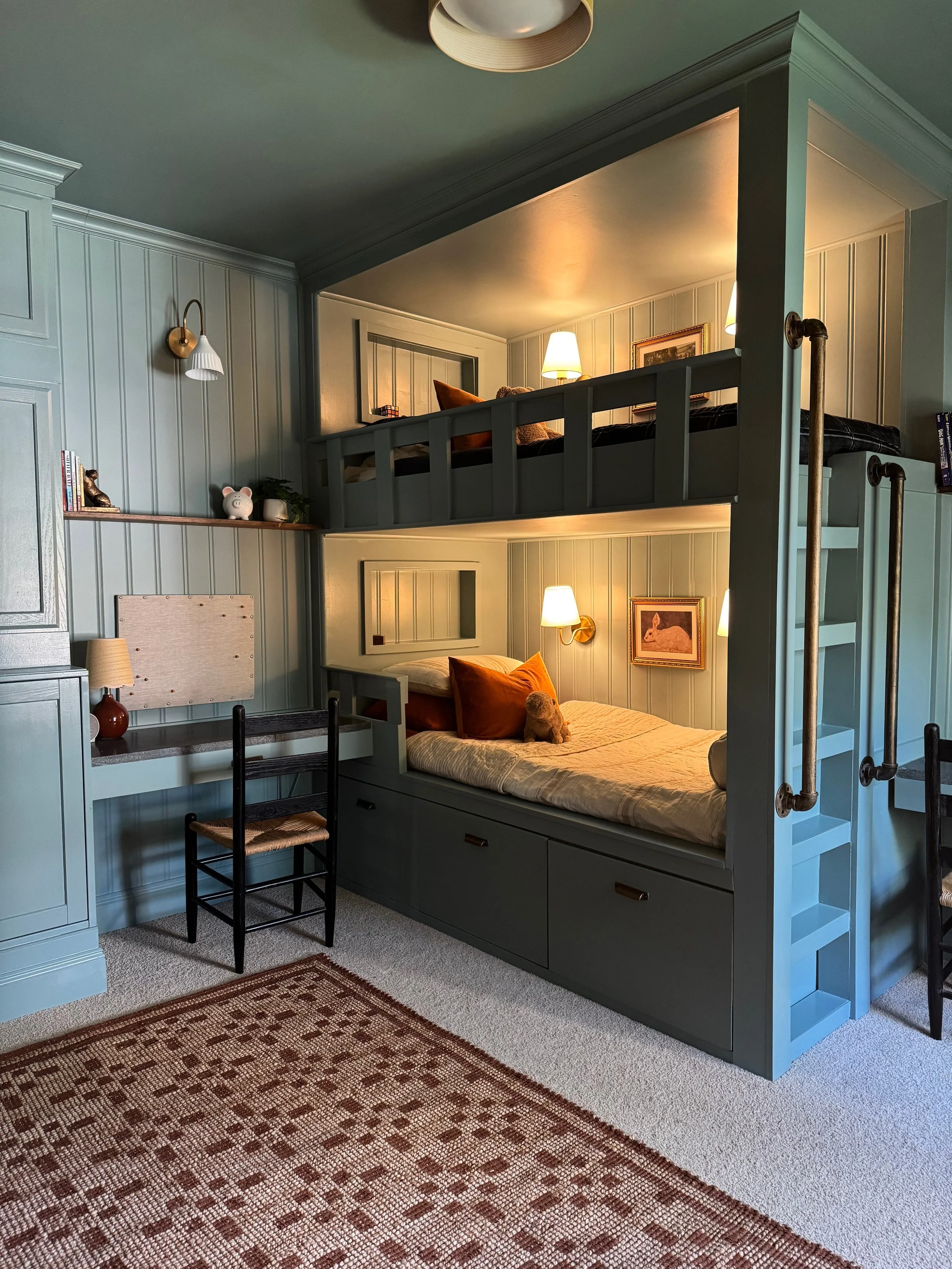 A cozy multi-level bedroom with a single bed on the lower level and a lofted bed above, both with warm lighting and decorated with pillows and stuffed animals. The room has light-green paneled walls, built-in storage drawers under the lower bed, and a small desk and chair. A decorative rug is on the carpeted floor.