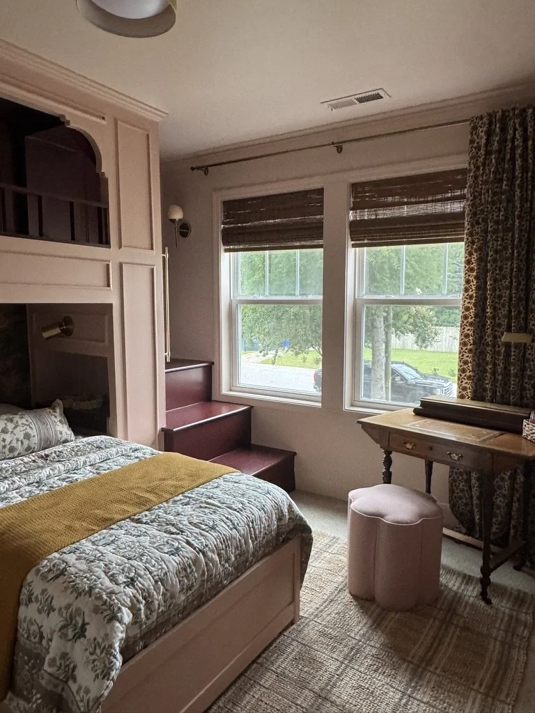 A cozy bedroom with a floral bedspread, a mustard-colored throw, and a window with dark blinds and a view of green trees outside. There is a small stairs by the window, a vintage-style desk with a rolled-up rug on top, a pink round ottoman, and a beige area rug.