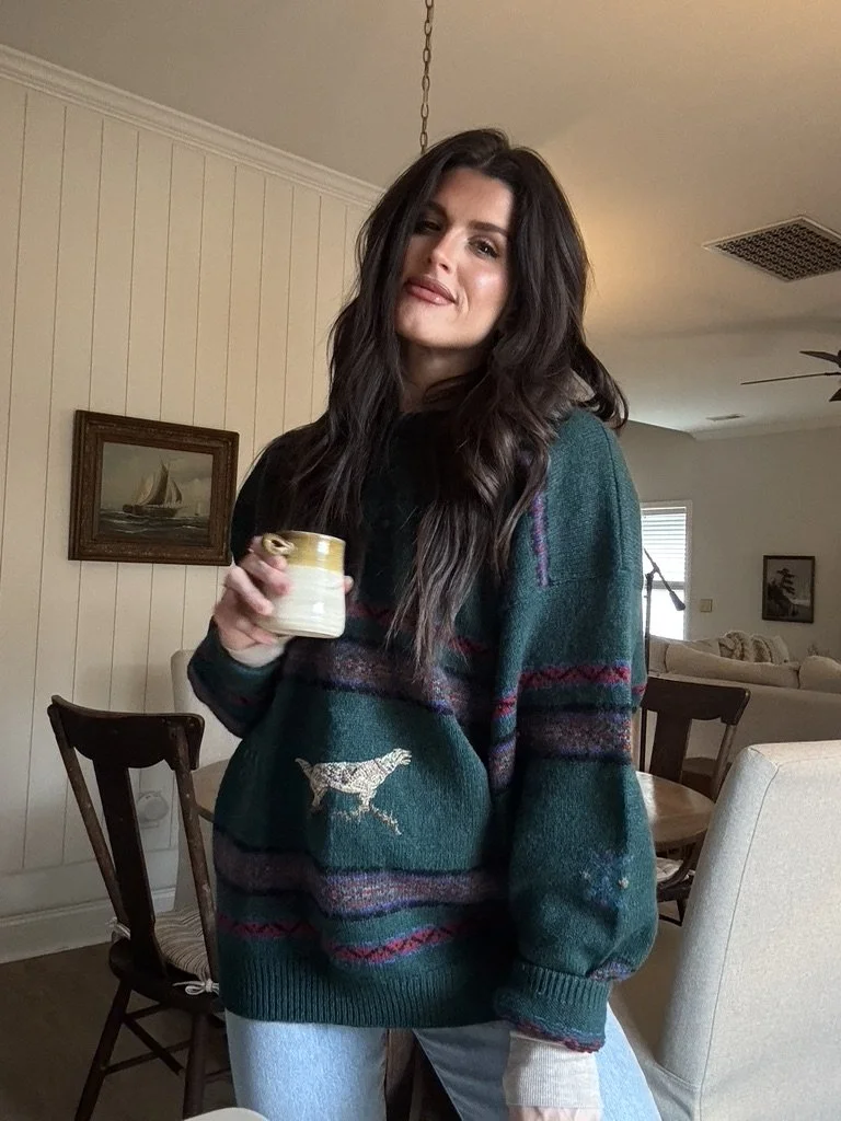 A woman with long dark hair holding a mug, standing in a cozy living room, wearing a green sweater with a dog design and colorful pattern.