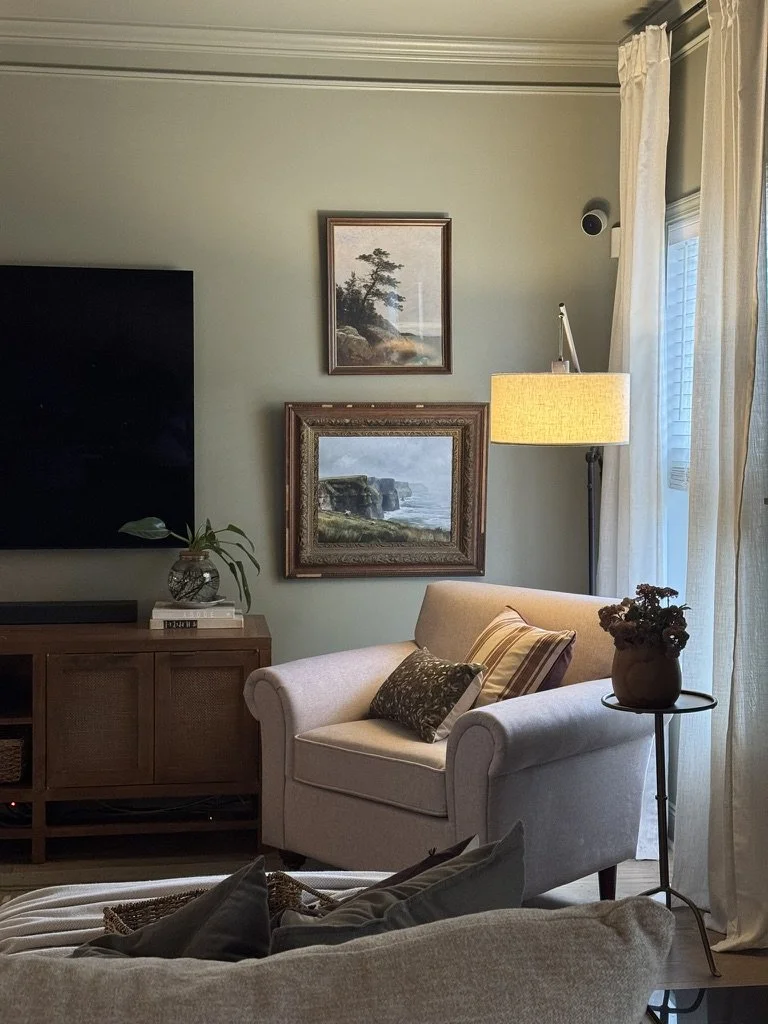 Cozy living room with a beige armchair, decorative pillows, a side table with a vase of dried flowers, a tall floor lamp, and a wall with two landscape paintings and a TV.