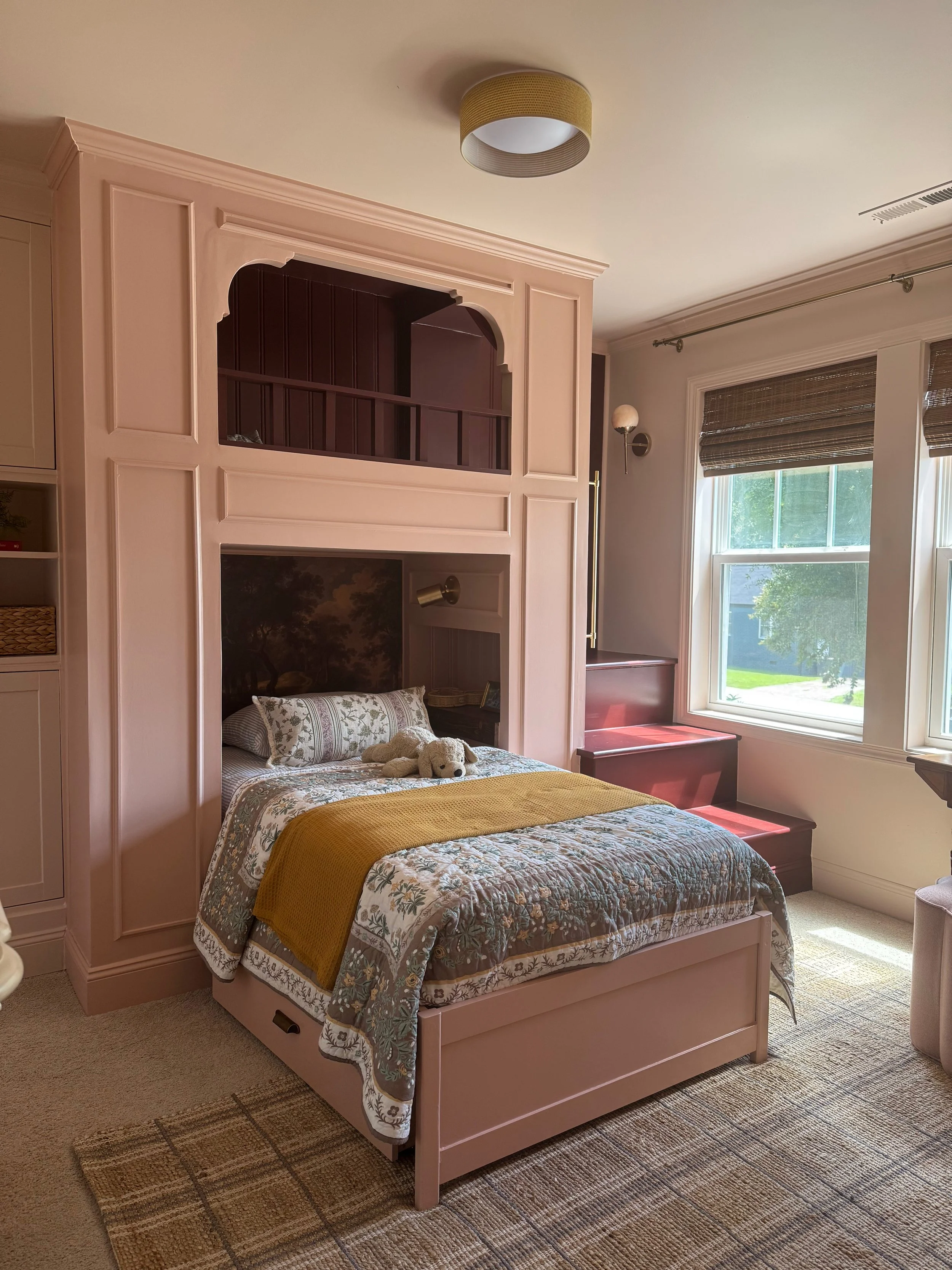 A pink bed with a floral patterned bedspread, a yellow blanket, two pillows, and a stuffed dog on it. The bed has a headboard with a mural of trees. Above the bed, there is a matching pink cabinet. To the right, there are two large windows with brown woven blinds, and a built-in red wooden staircase with a seat. The room has beige carpet and a modern ceiling light.