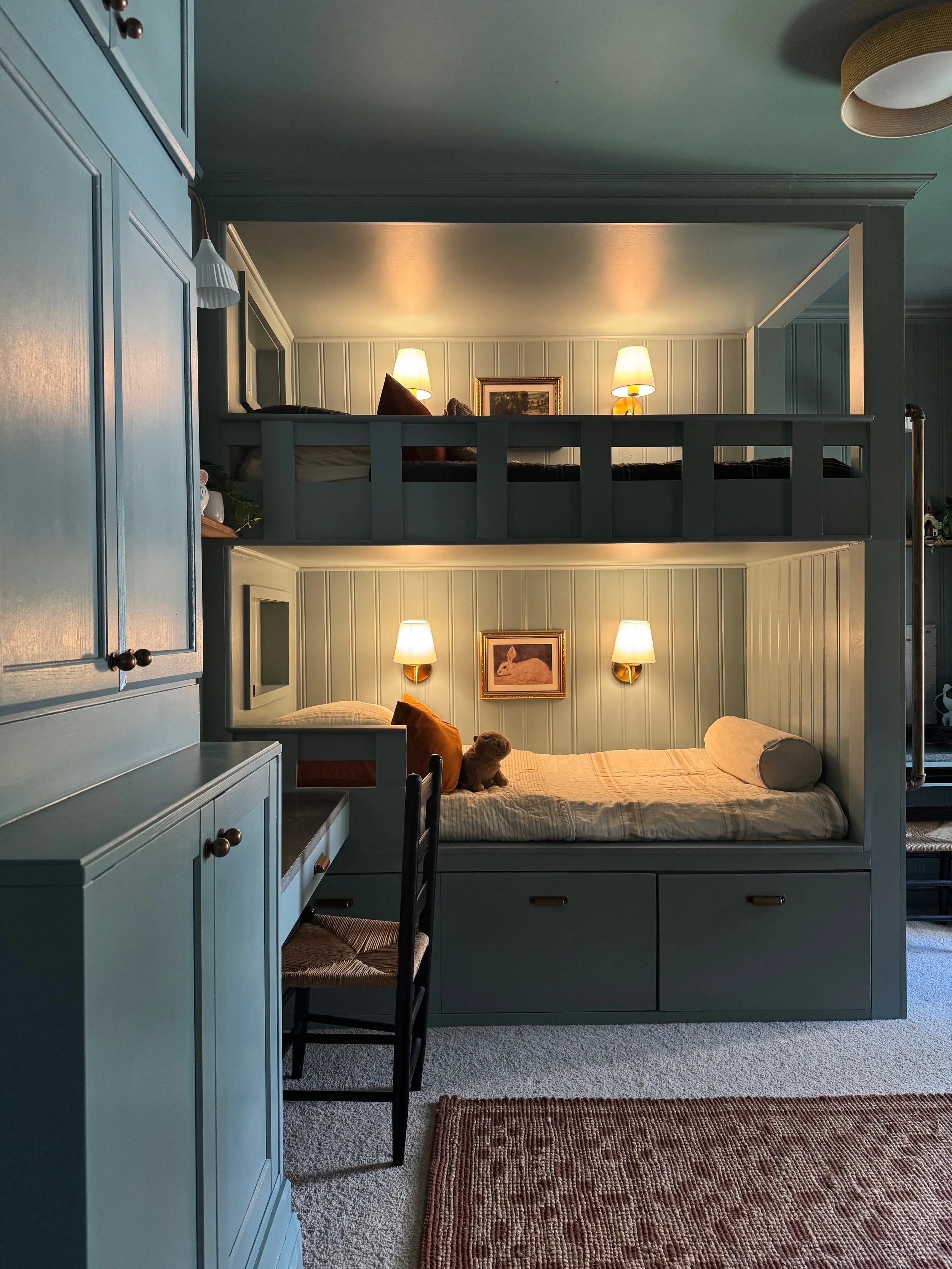 Bunk beds with a lower bed and lofted upper bed in a cozy room, with blue-gray paneling and small lamps, a framed picture, stuffed toy, and cushions.