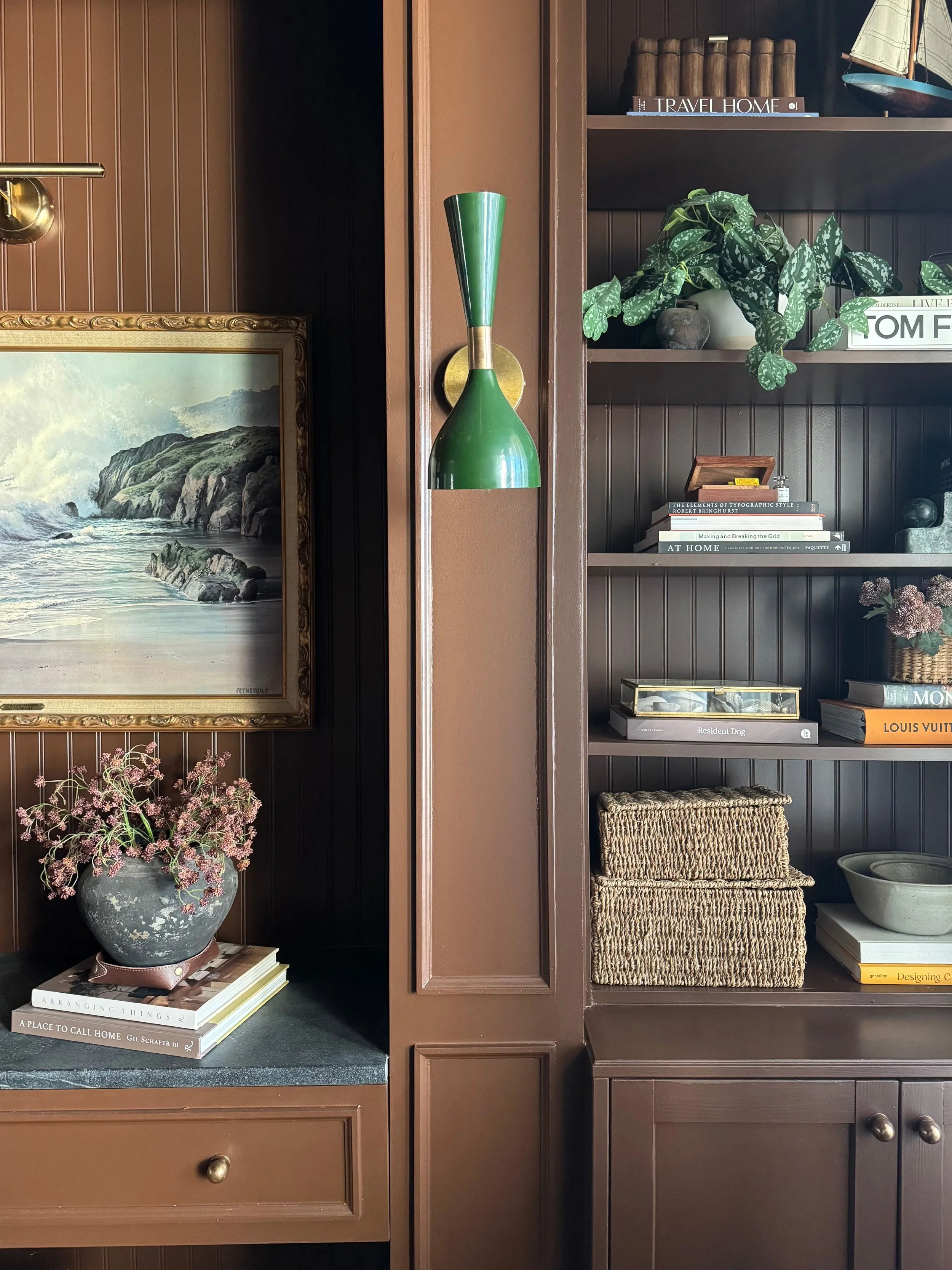 Interior view of a bookshelf and a corner with decorative items, including a painting, a potted plant, books, and a vintage wall-mounted lamp.