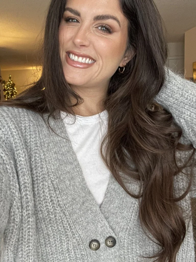 A woman with long brown hair, smiling and showing her teeth, wearing a white top and a gray knit cardigan, indoors with a warm lighting and a blurred background.