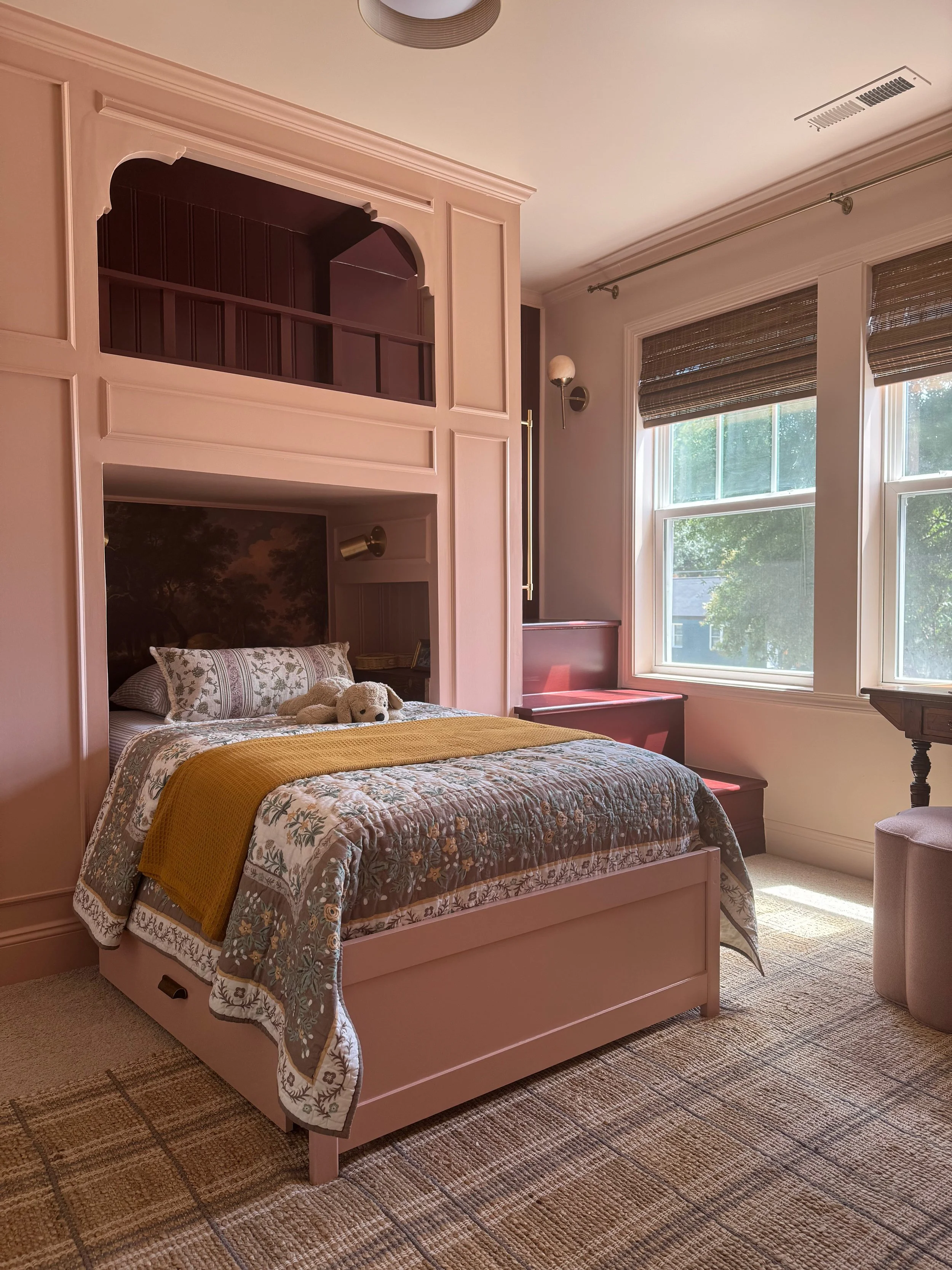 A cozy bedroom with a pink bed frame, floral bedding, and a stuffed animal. Natural light streams in through large windows with brown blinds. There are built-in shelves above the bed, wall-mounted lights, and a sitting area by the window.