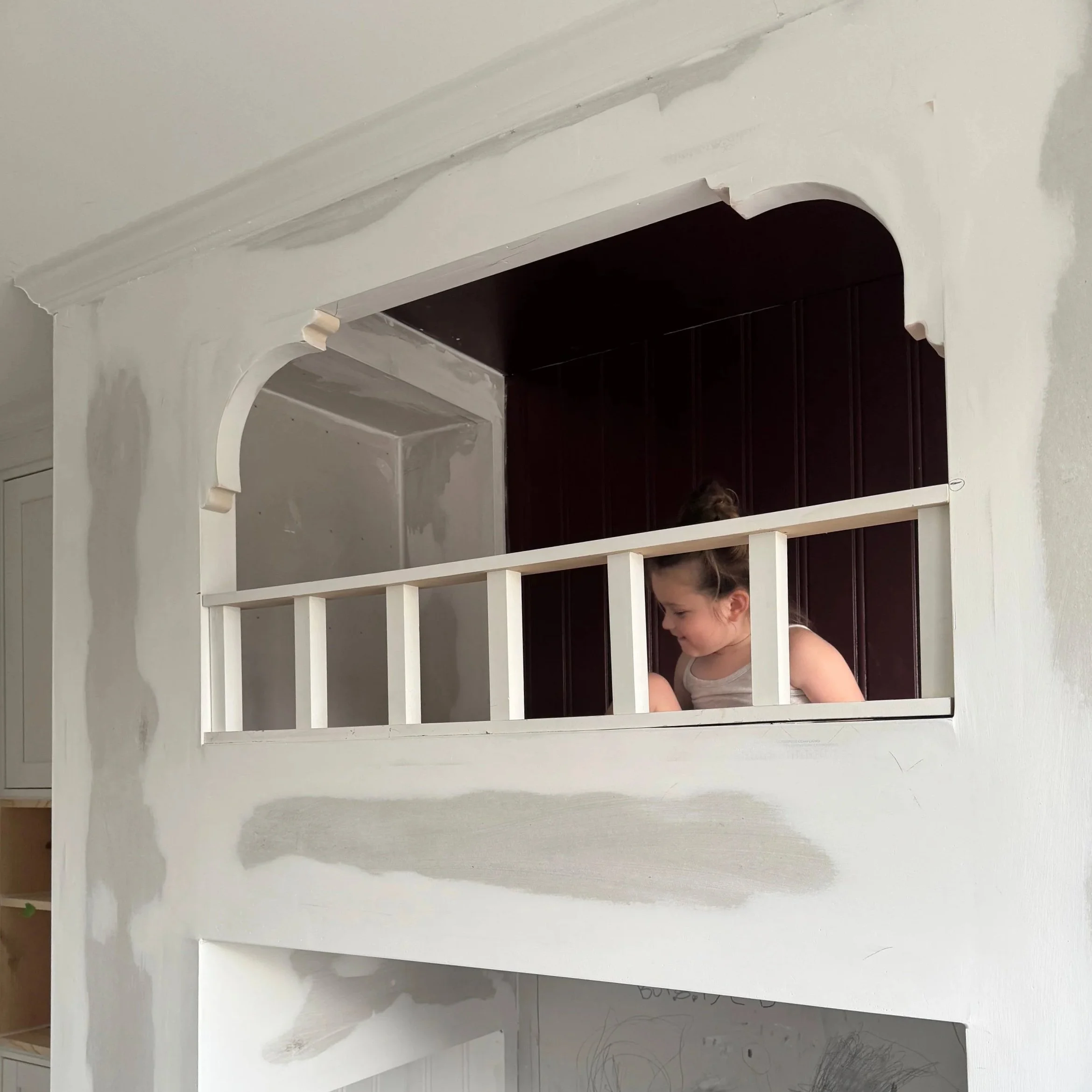A young girl leaning on a white wooden railing in a partially constructed or renovated wall with some patches of drywall and a dark interior space behind her.