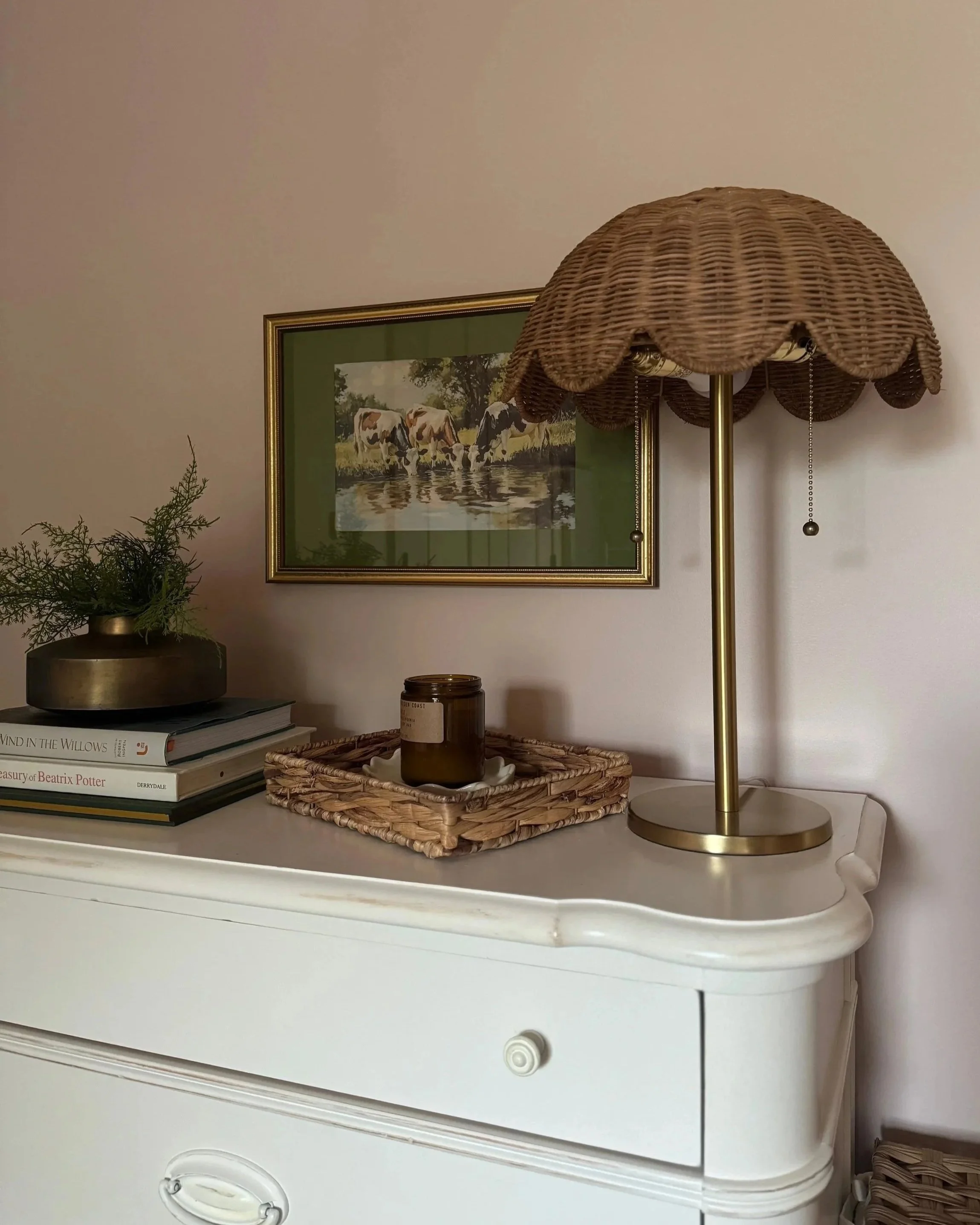 A white dresser with a wicker basket tray holding a small candle, a brass table lamp with a woven shade, a stack of books, a glass vase with greenery, and a framed picture of cows on a fence hanging on the wall behind.