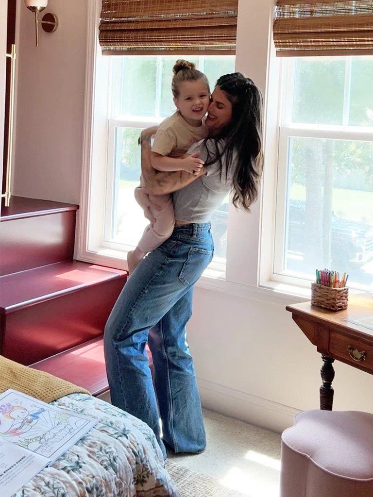 A woman holding a young girl in a sunlit room with two large windows, a wooden desk with colored pencils, and a bed with children's coloring pages.
