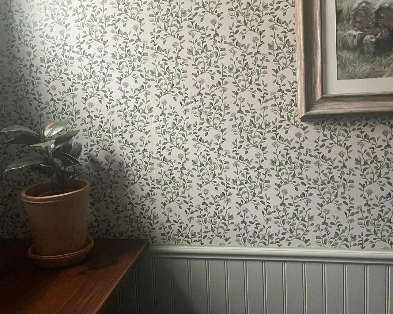 A potted plant with dark green leaves rests on a wooden table in front of a wall covered with cream-colored wallpaper decorated with a leafy vine pattern. A framed picture hangs partially on the right side of the image.