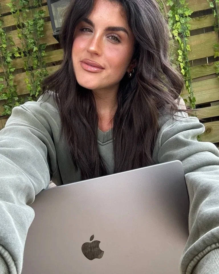 Young woman with long dark hair taking a selfie outdoors, holding a MacBook laptop, with a wooden fence and green plants in the background.