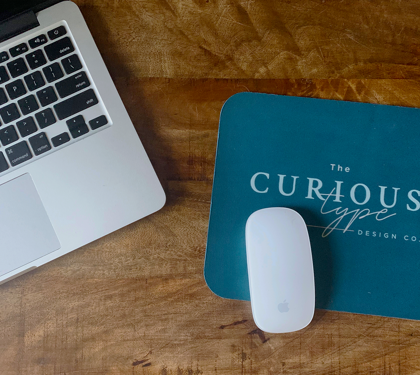 Laptop keyboard and trackpad, white Apple Magic Mouse, and a blue folder with the text 'The Curious Type' on a wooden desk.