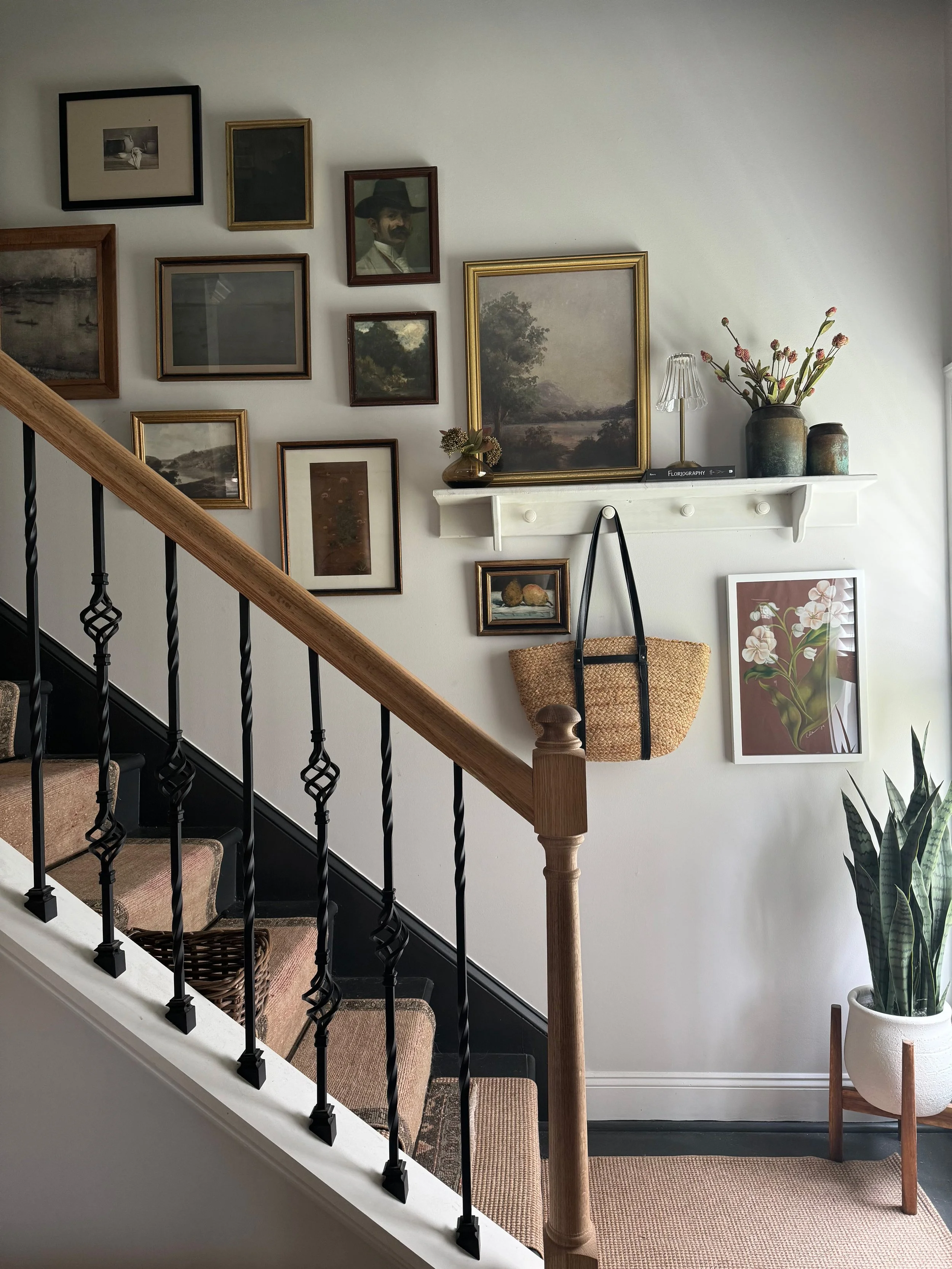 Interior view of a staircase with a wall decorated with various framed pictures, paintings, and photographs, including a landscape and a portrait. There's a white shelf with vases, a small lamp, and a book. A woven tote hangs from a hook below the shelf. A large framed botanical print is on the wall, and a tall potted snake plant stands on the floor near the stairs.
