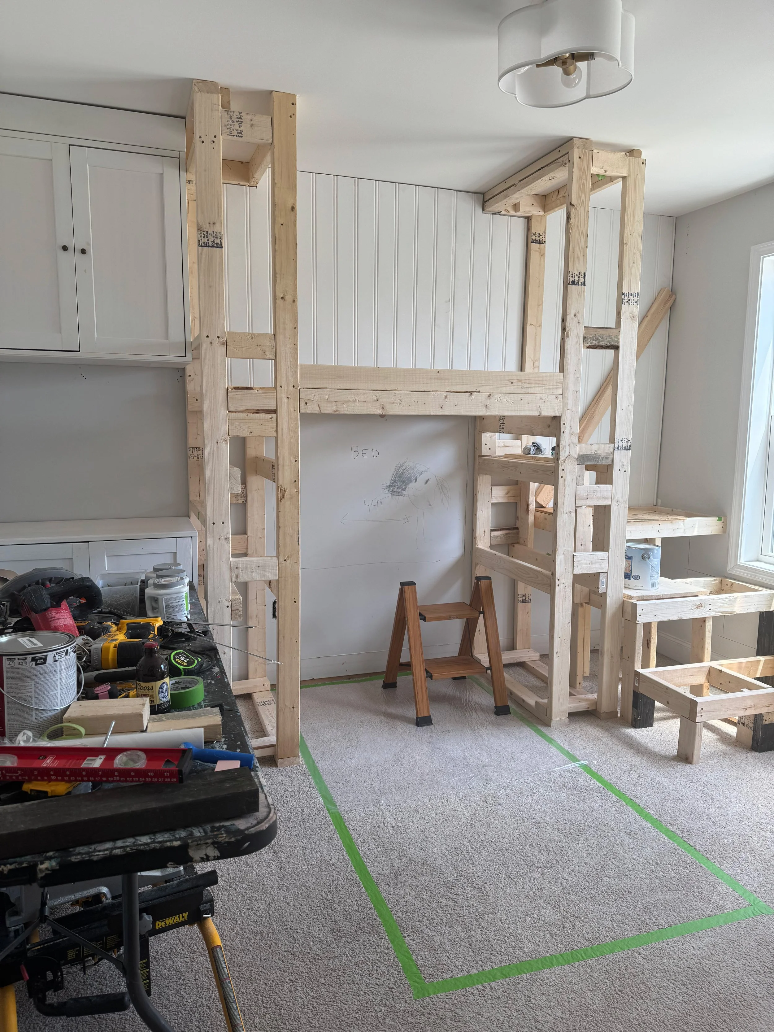 Unfinished room with a wooden loft bed frame under construction, a small step stool, tools and supplies on a work table, and a marked outline on the carpet for the bed area.