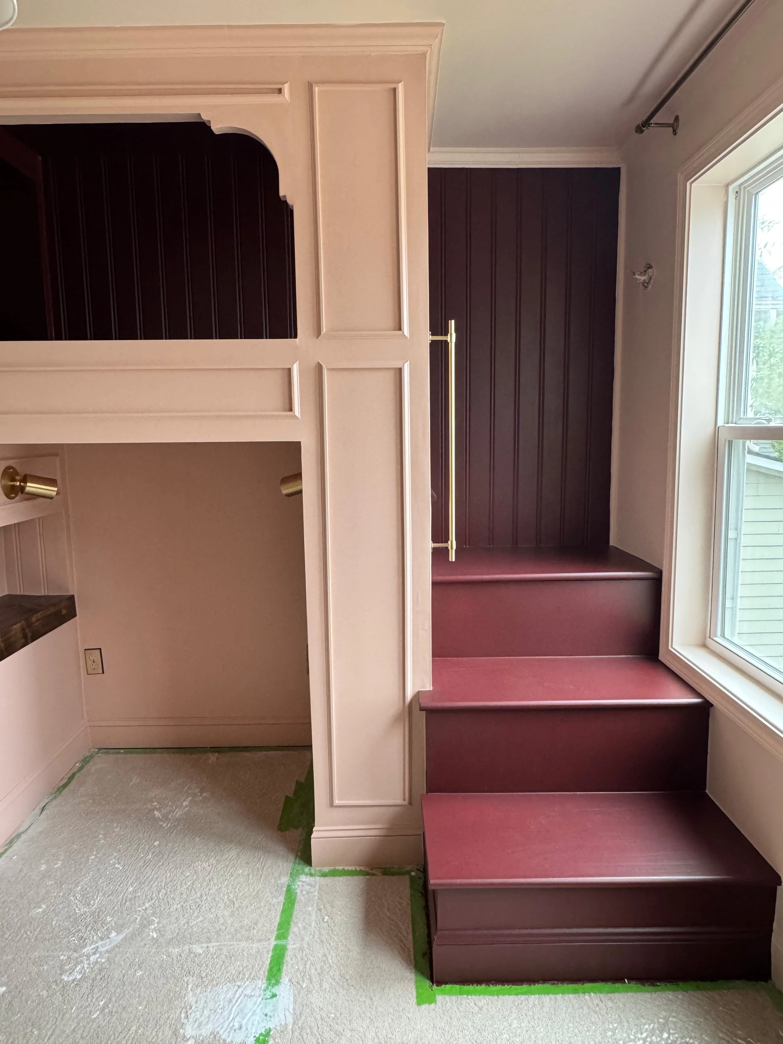 Interior of a room with built-in pink and dark purple woodwork, including stairs and a bench, next to a large window with white trim.