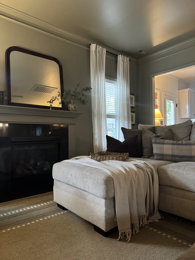 Living room with a beige sectional sofa, black, gray, and plaid pillows, a beige blanket and throw on the ottoman, a fireplace with a mirror and decorative vase on mantle, and windows with sheer white curtains letting in natural light.