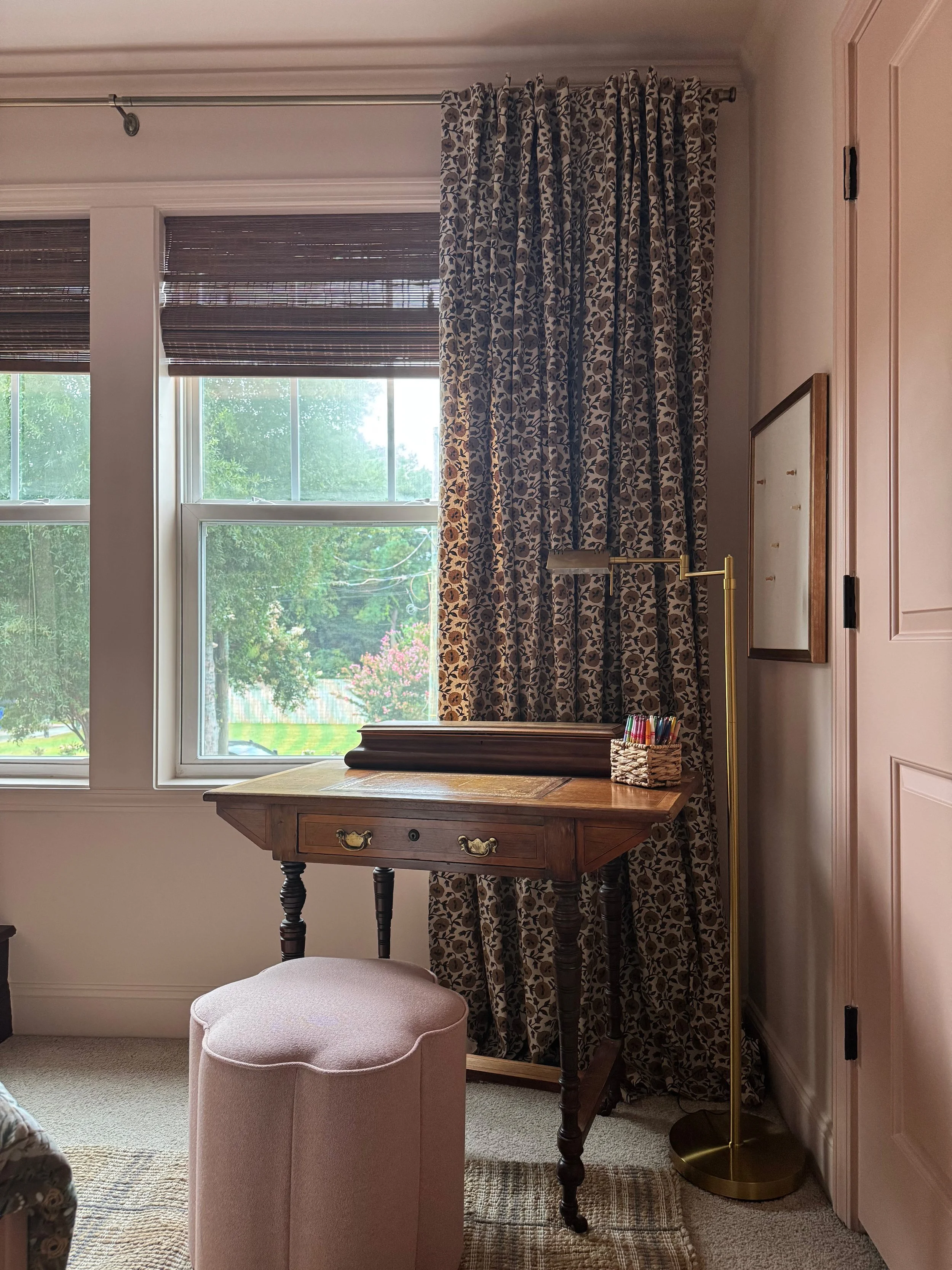 A corner of a room with a window and a floral curtain, an adjustable floor lamp, a vintage wooden desk with brass handles, a small woven basket filled with markers, a pink upholstered stool, and a patterned rug.