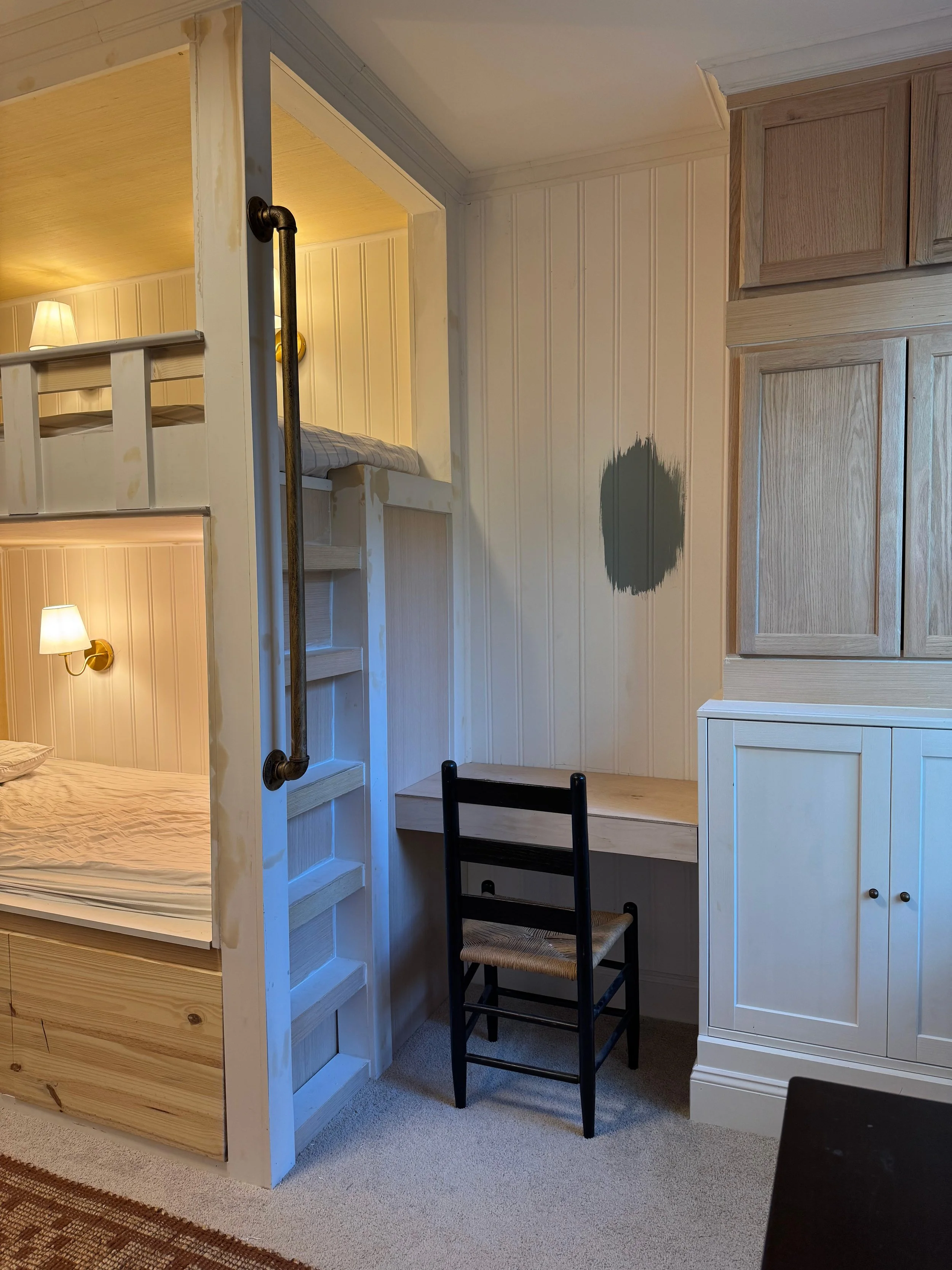 Interior of a room with a bunk bed, small desk, black chair, and wooden cabinets; patch of unpainted wall on the white wooden wall.