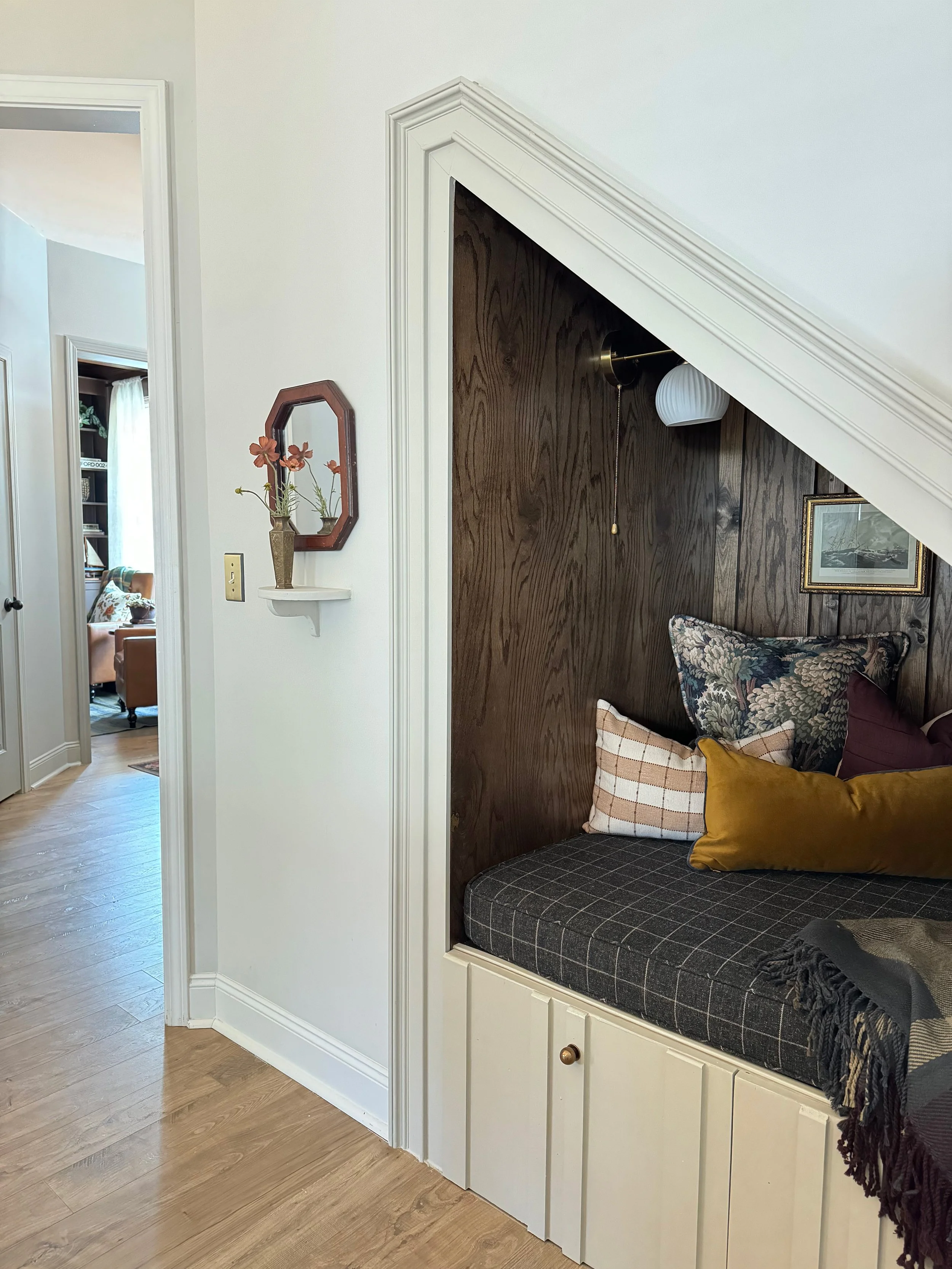 Corner nook with built-in seating under a staircase, decorated with pillows, with wood paneling and artwork.
