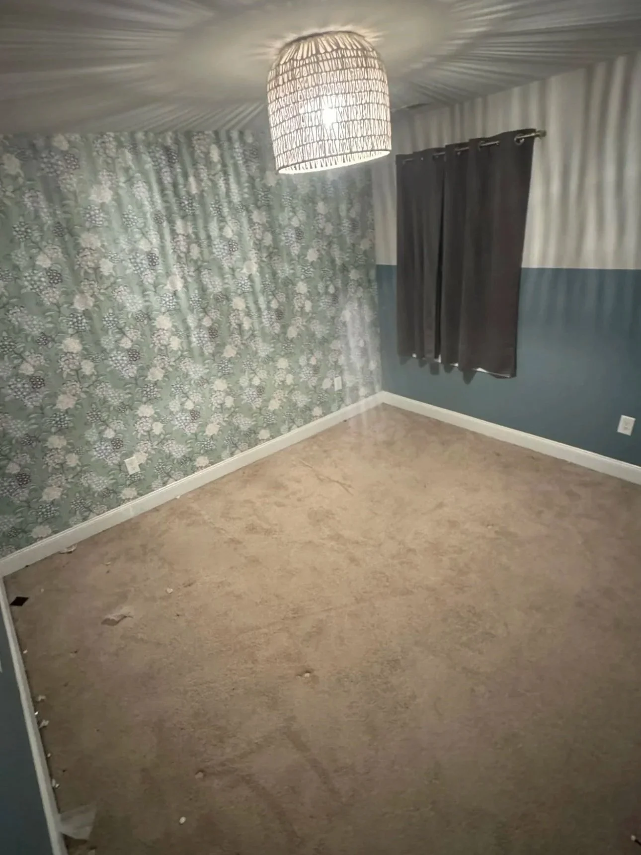 Empty room with patterned wallpaper on one wall, dark curtains over a window, and beige carpet flooring.
