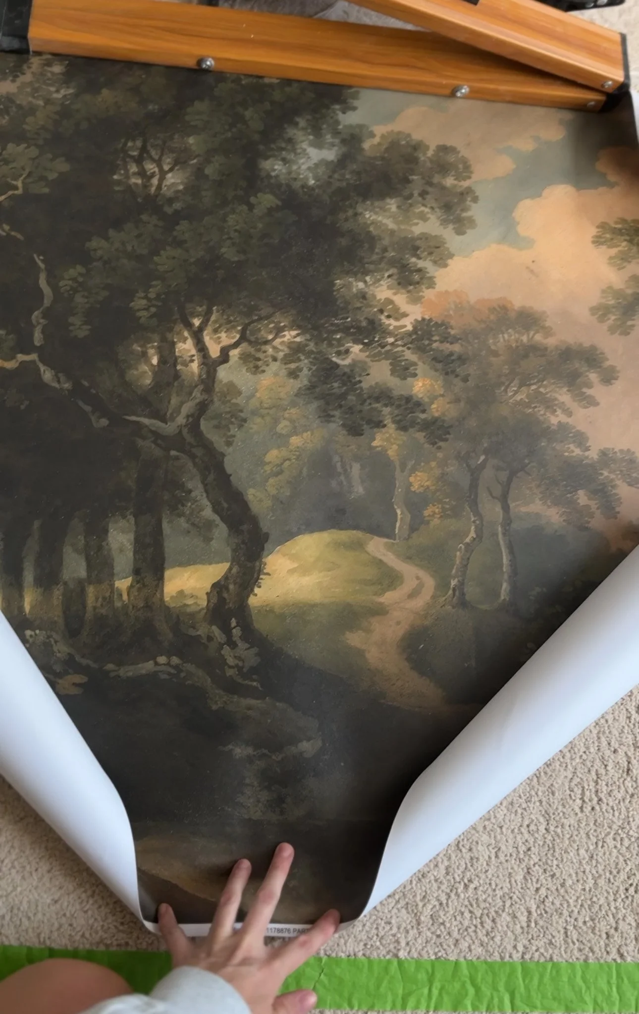 A framed landscape painting or print of a forest scene with tall, twisting trees, a dirt path, and a soft, cloudy sky. The artwork is partially curled, with a child's hand holding the bottom edge.