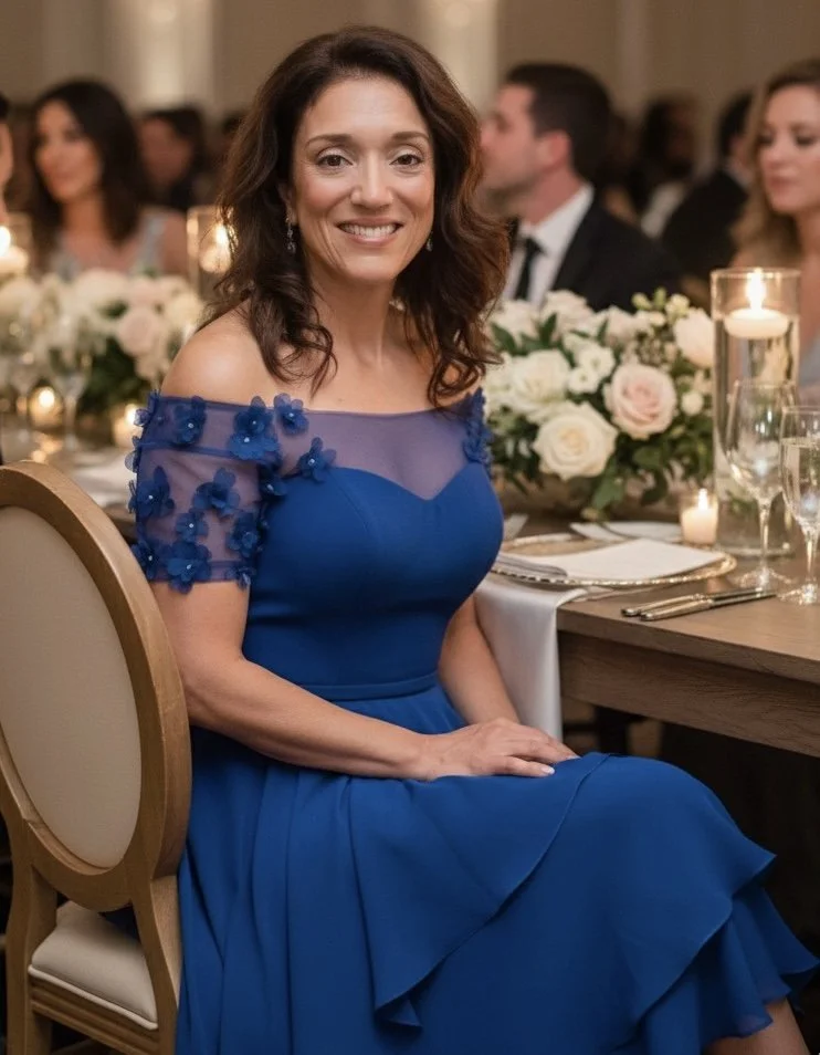 A woman wearing a blue dress with floral embellishments sitting at a formal dinner event with floral centerpieces and candles.