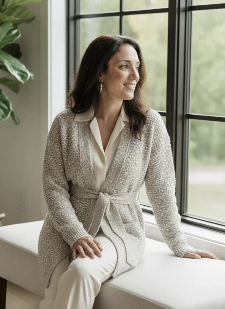 A woman with dark brown hair sitting on a white window seat, looking out the window with a smile, wearing a beige cardigan, cream blouse, and cream pants, with a large green plant to her left and a nature view outside.