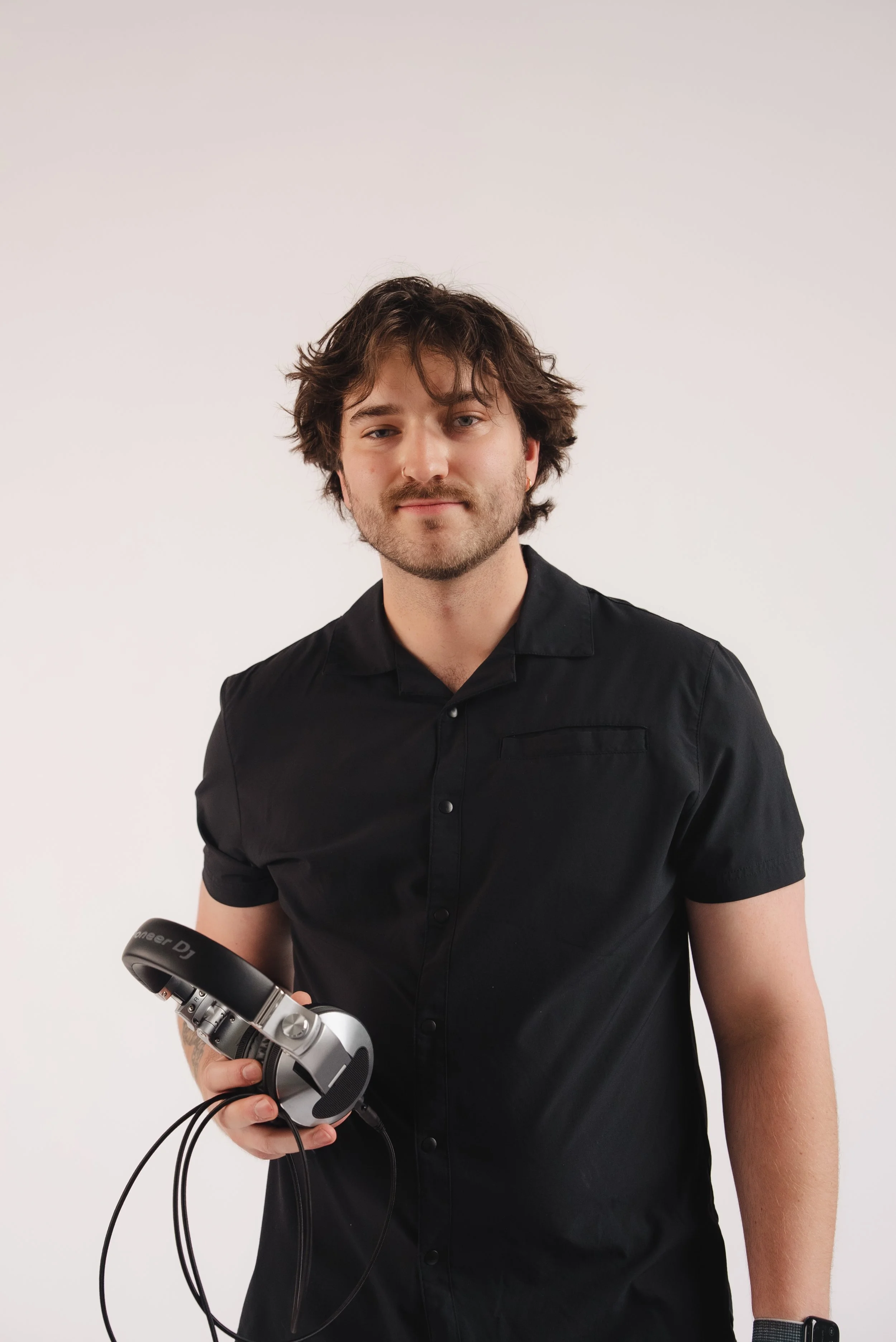 A young man with dark hair and a beard, wearing a black shirt, standing against a plain white background, holding a pair of headphones in his right hand.