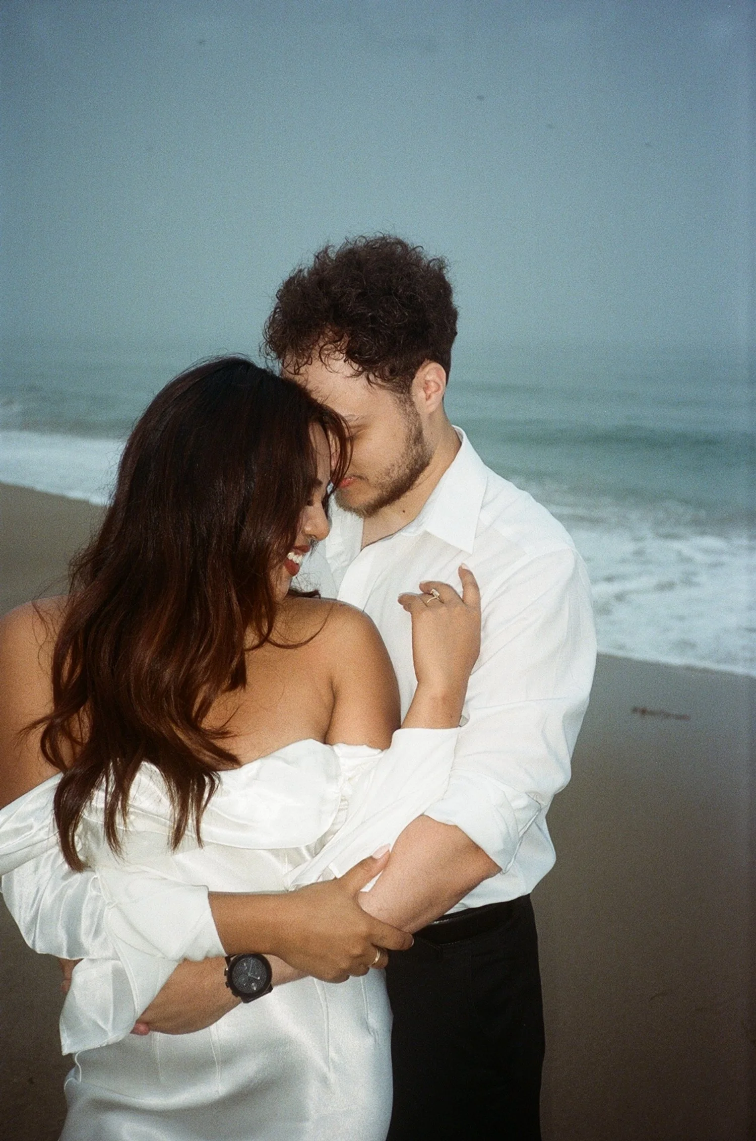 A couple embracing on the beach, with the ocean and waves in the background, during what appears to be early evening.
