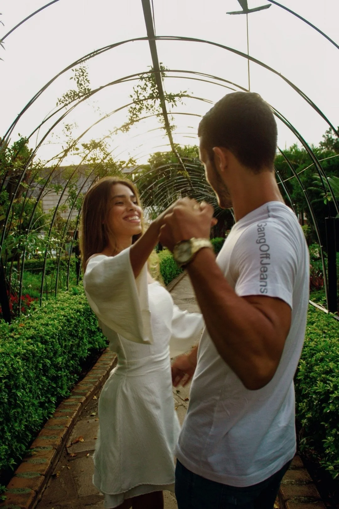 A couple dancing outdoors in a garden with green plants and arched metal structures overhead during sunset.