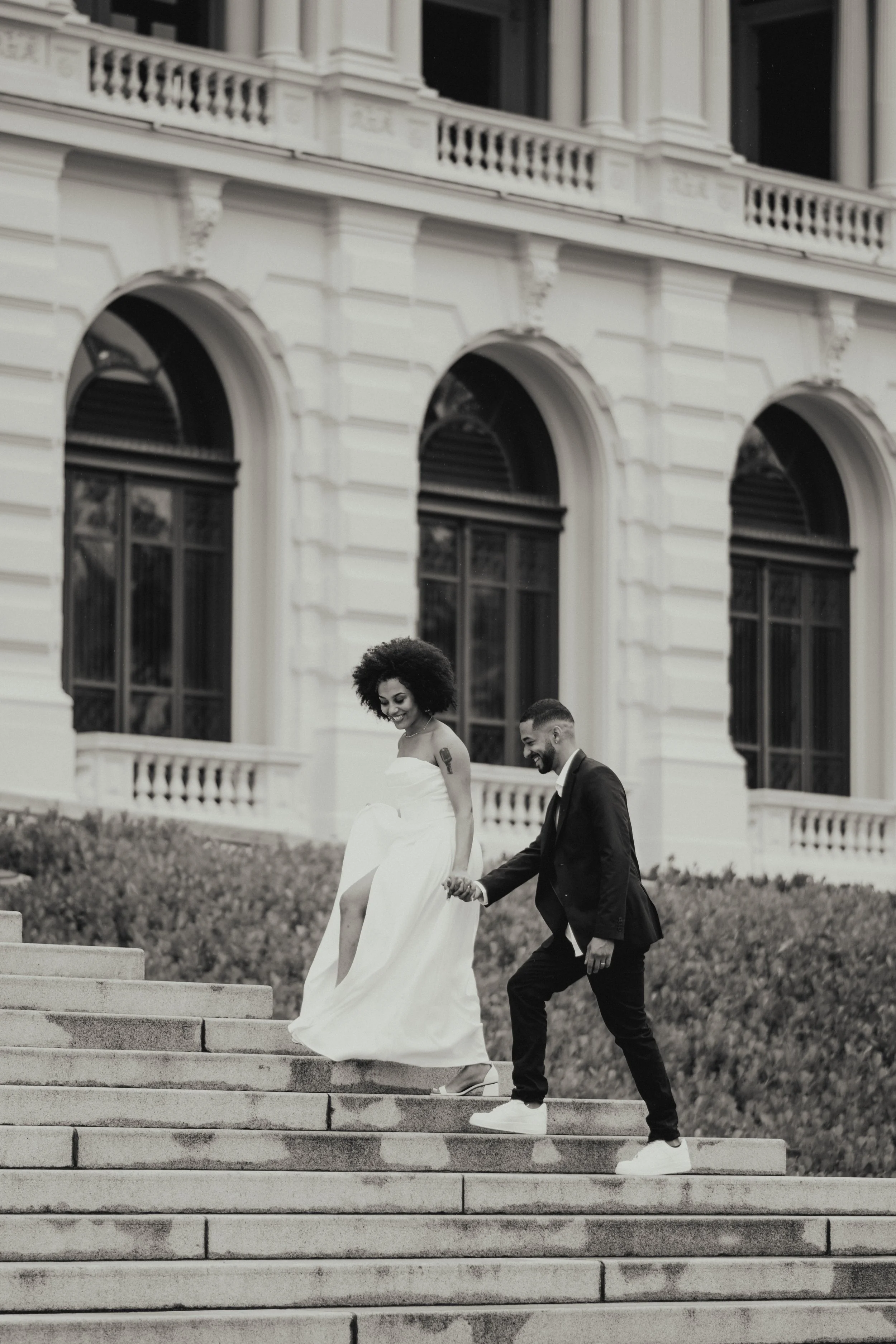 A couple dressed in wedding attire walking down outdoor stairs in front of a historic building with arched windows.