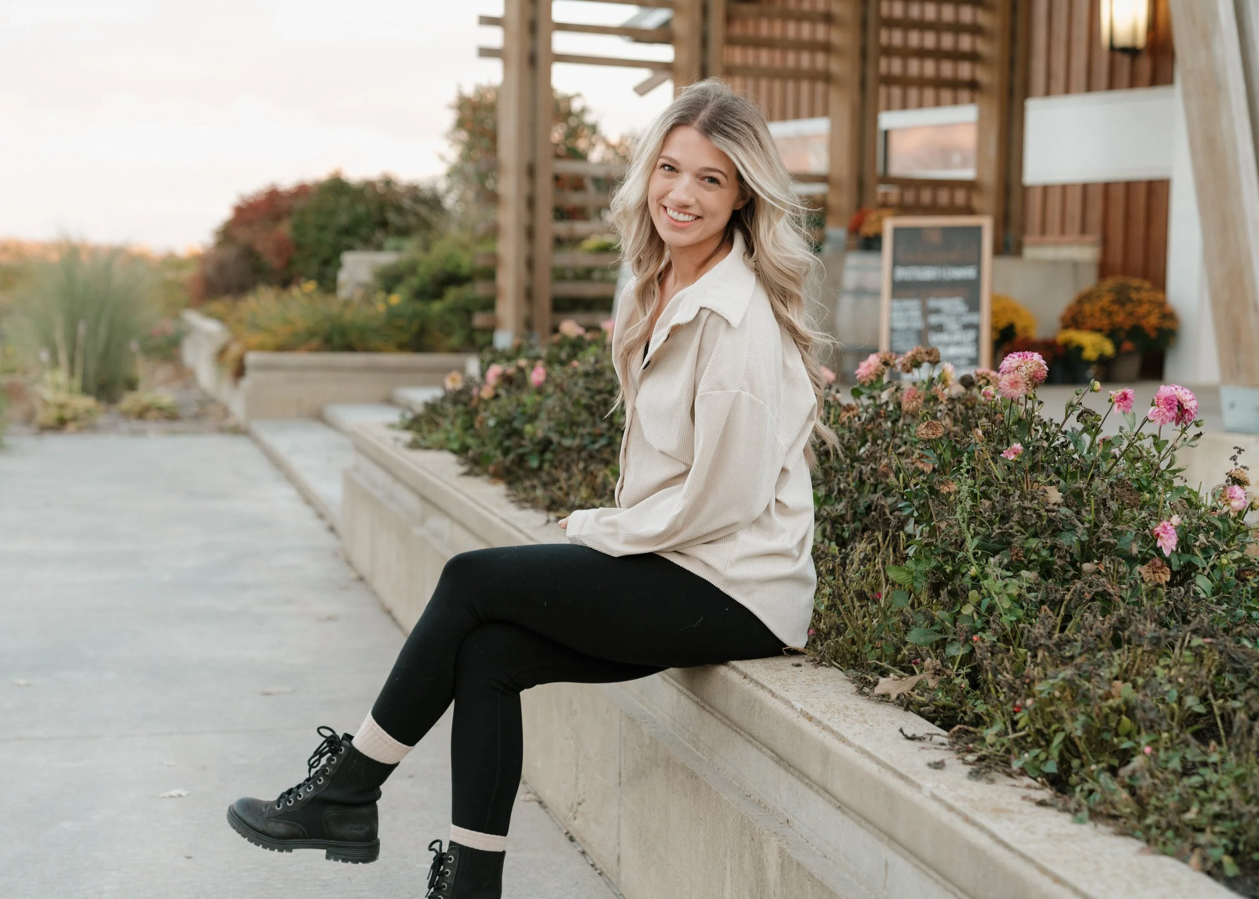 A young woman with long blonde hair, smiling and sitting on a concrete ledge beside a flower bed with pink and purple flowers. She is wearing a beige jacket, black pants, white socks, and black combat boots. Behind her, there is a wooden building and outdoor stairs.