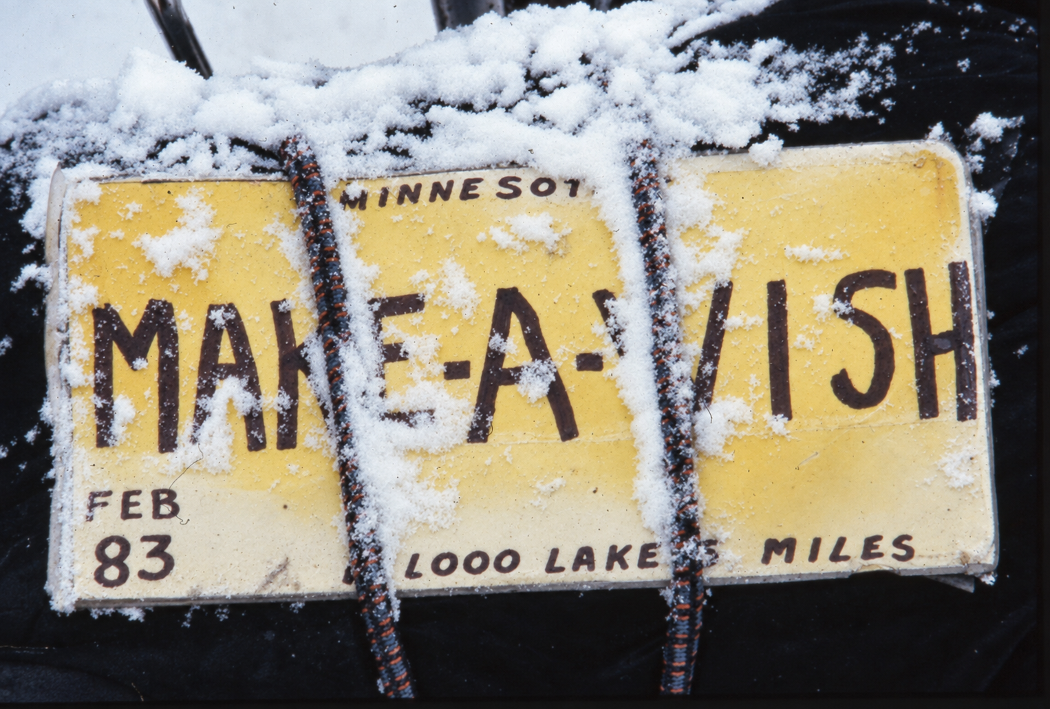A yellow Minnesota license plate covered in snow, with details indicating it's from February 1983, for 1000 Lake and miles.