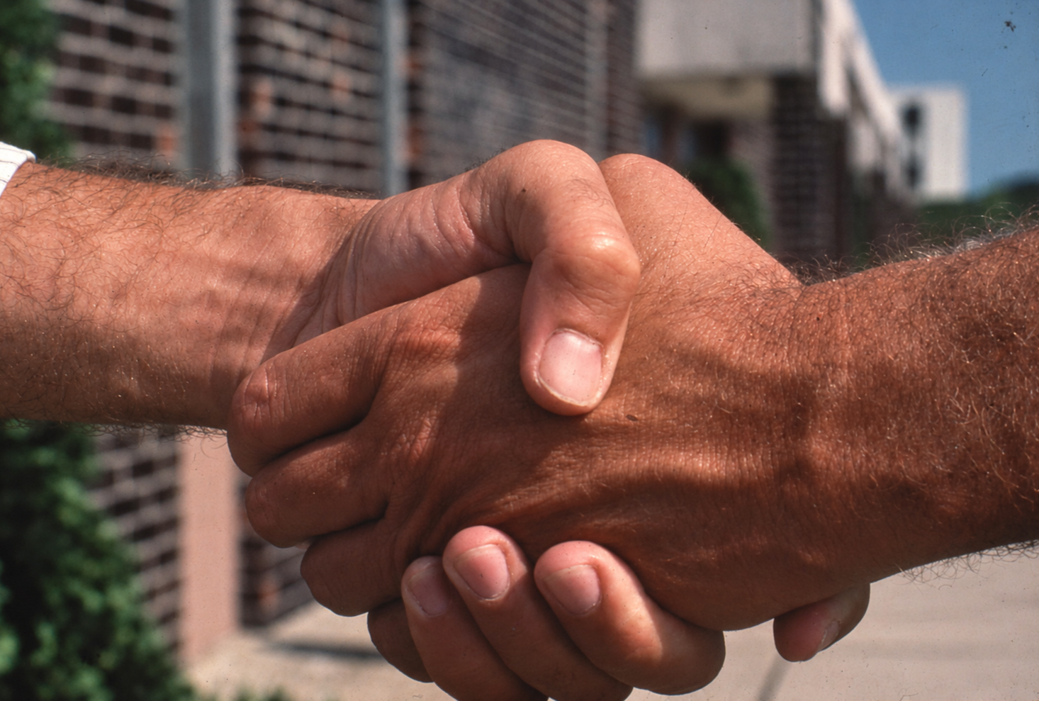 Close-up of two men shaking hands outdoors in front of buildings.