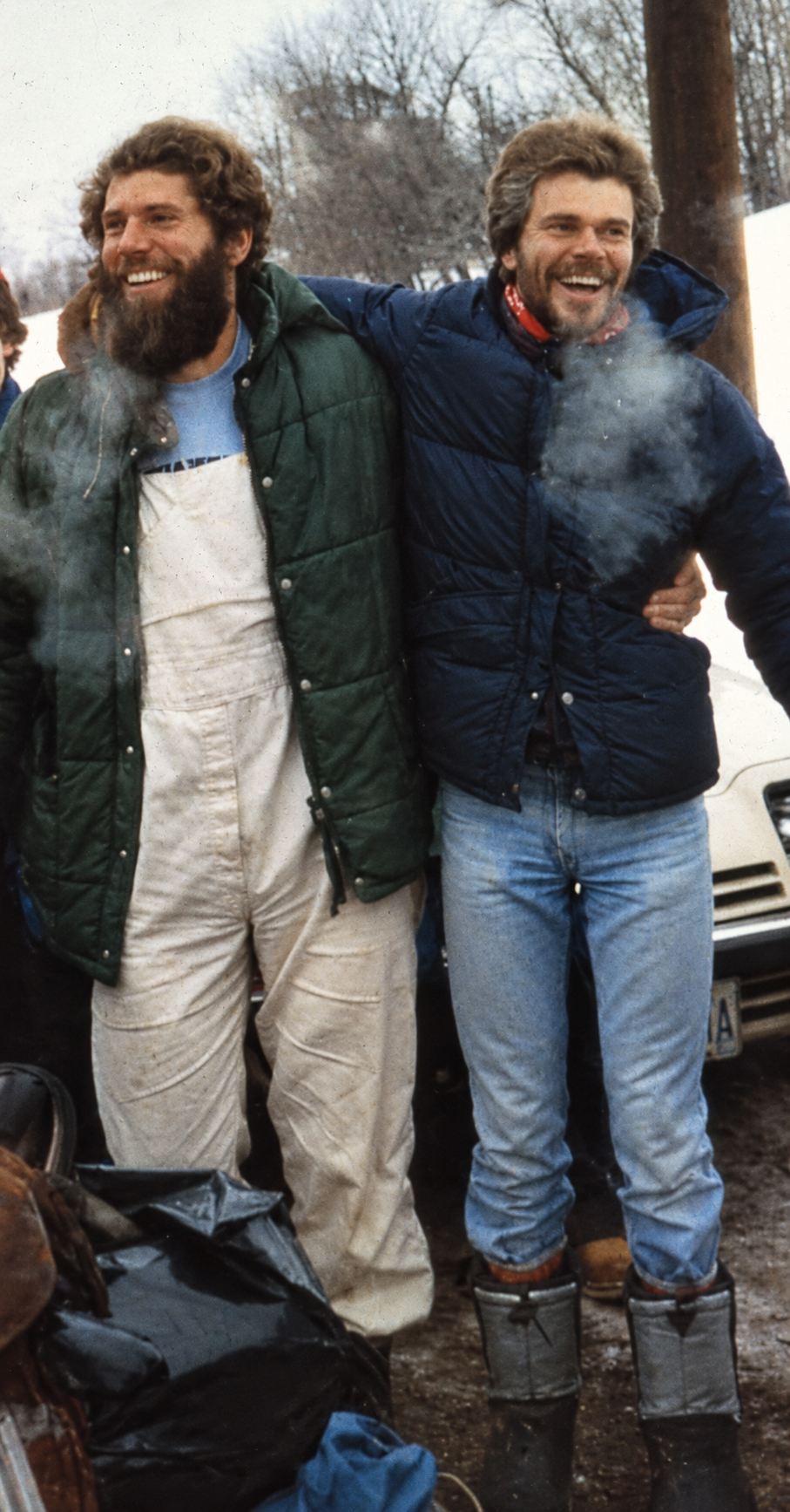 Two smiling men with beards, wearing warm jackets and jeans, standing outdoors in a winter setting with snow, trees, and a vehicle in the background, enjoying each other's company.