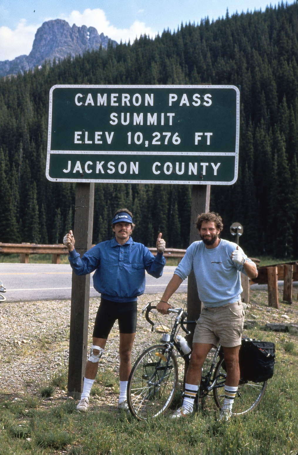 Two men standing next to a bicycle under a sign that reads 'Cameron Pass Summit Elev 10,276 ft Jackson County'. They are giving thumbs-up signs, and there are mountains and a forest in the background.